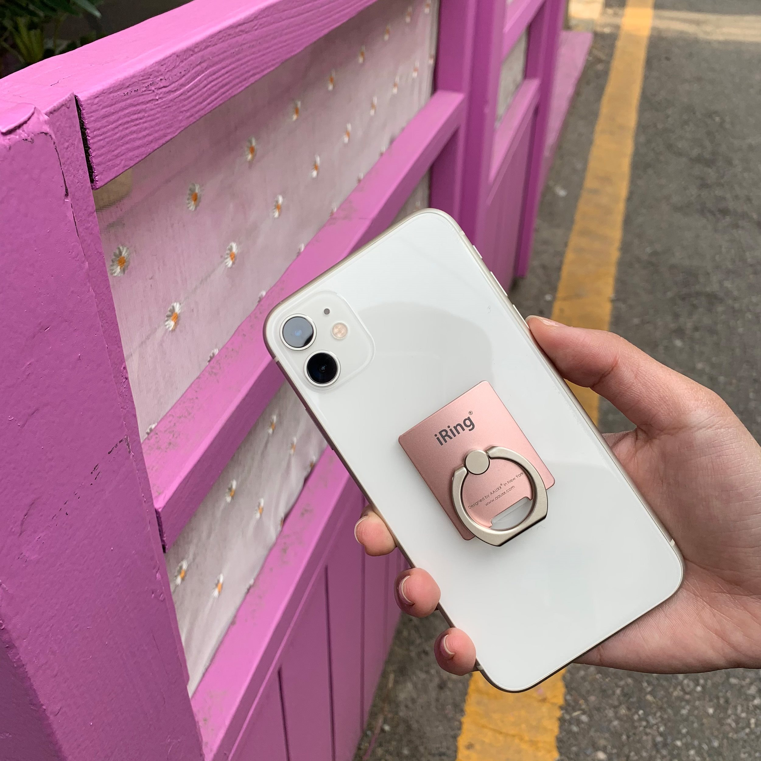 Weißes iPhone mit einem pinken Telefonringhalter auf einem pinken Zaunhintergrund. Hand hält das Telefon.