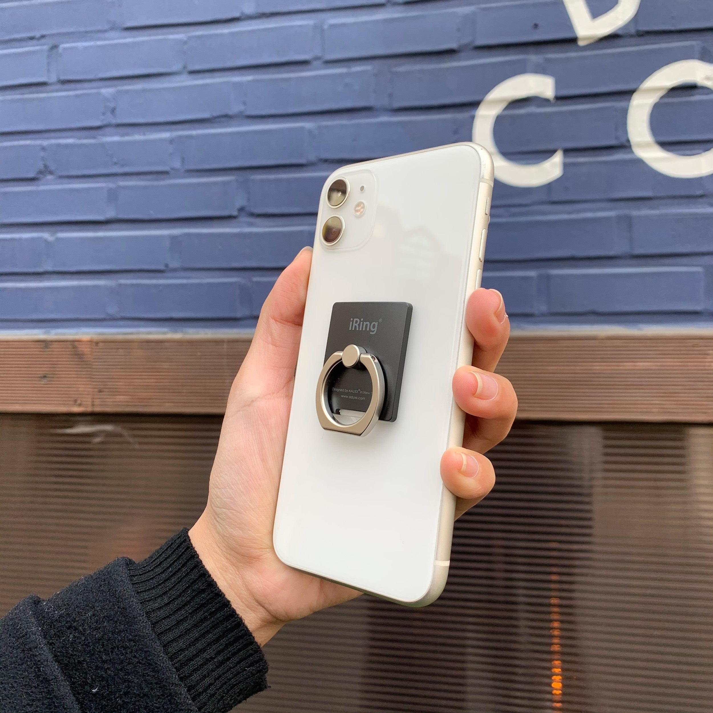 Eine Hand, die ein weißes Telefon mit einem daran befestigten schwarzen Ringhalter hält, vor einer Ziegelmauer.