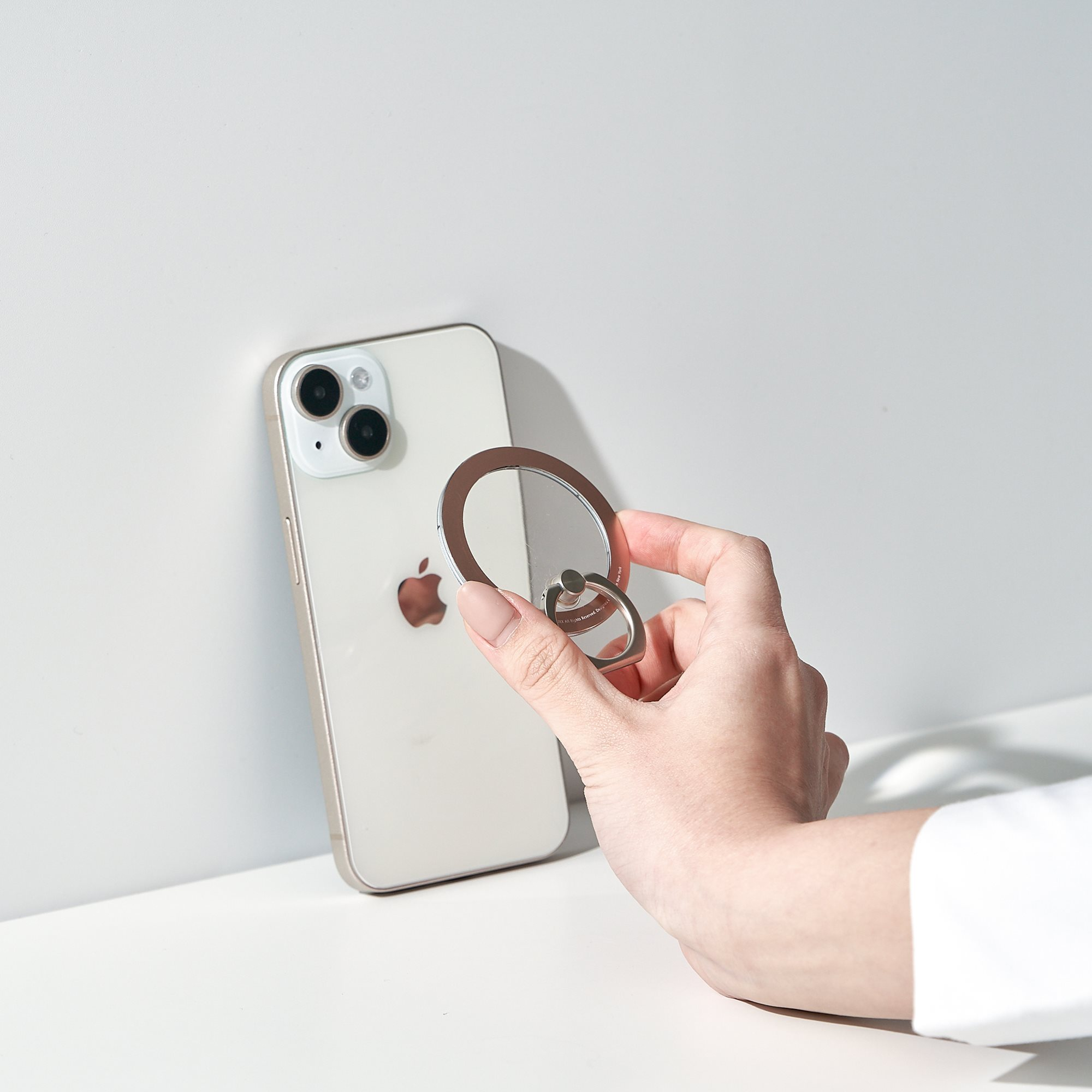 Eine Hand hält einen silbernen Telefonring, der an einem weißen iPhone befestigt ist, auf einer weißen Oberfläche.