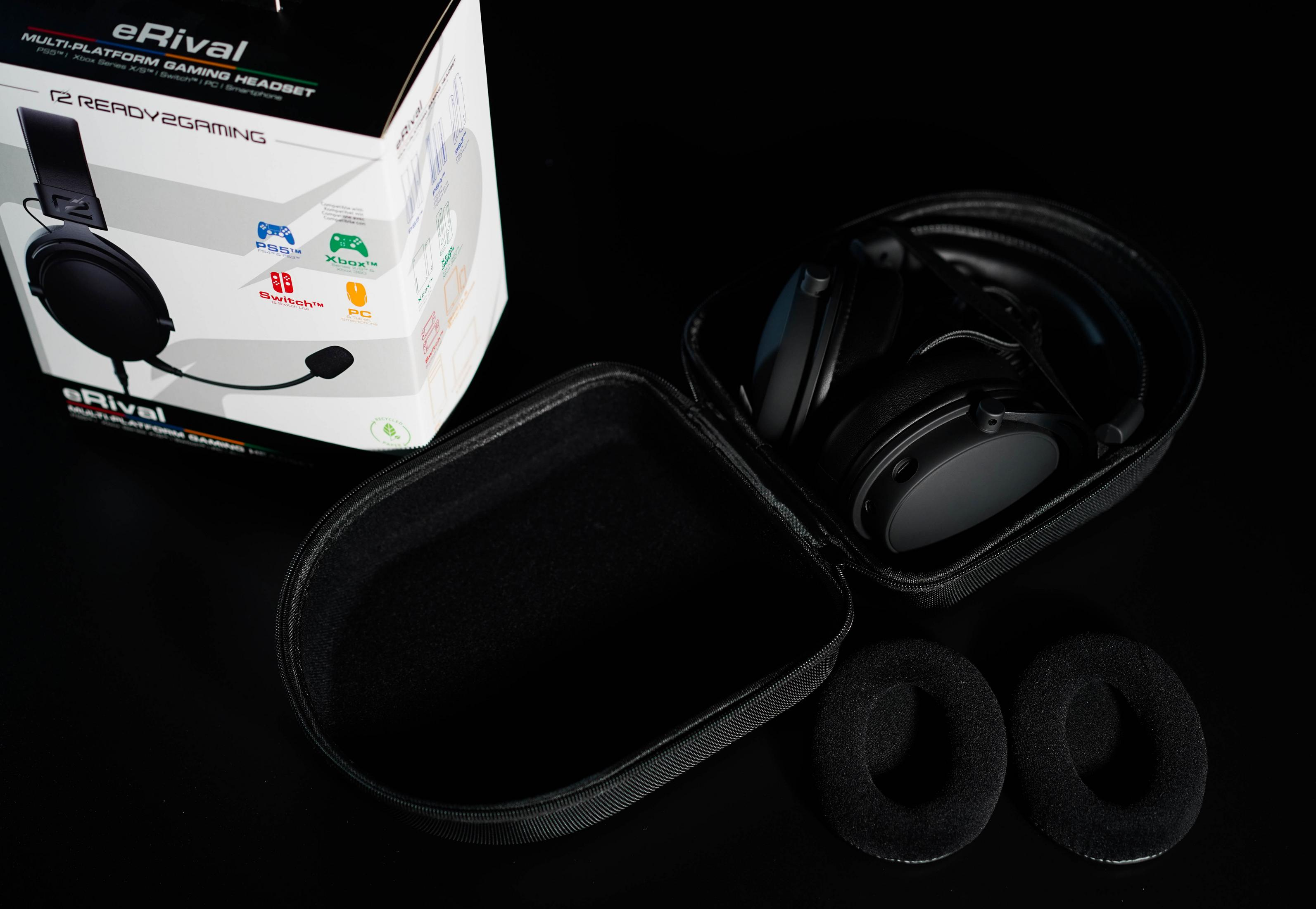 Schwarzes Gaming-Headset, Etui und Ohrpolster auf schwarzer Oberfläche. Box im Hintergrund. Alle Elemente gut beleuchtet.