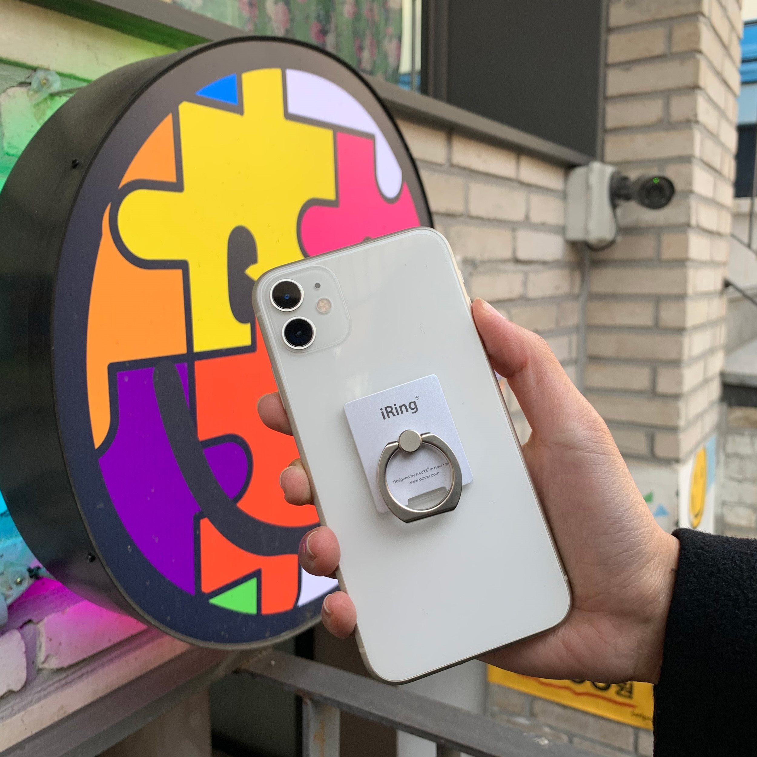 Hand hält ein weißes Telefon mit Ringgriff. Ein farbenfrohes Schild ist im Hintergrund.