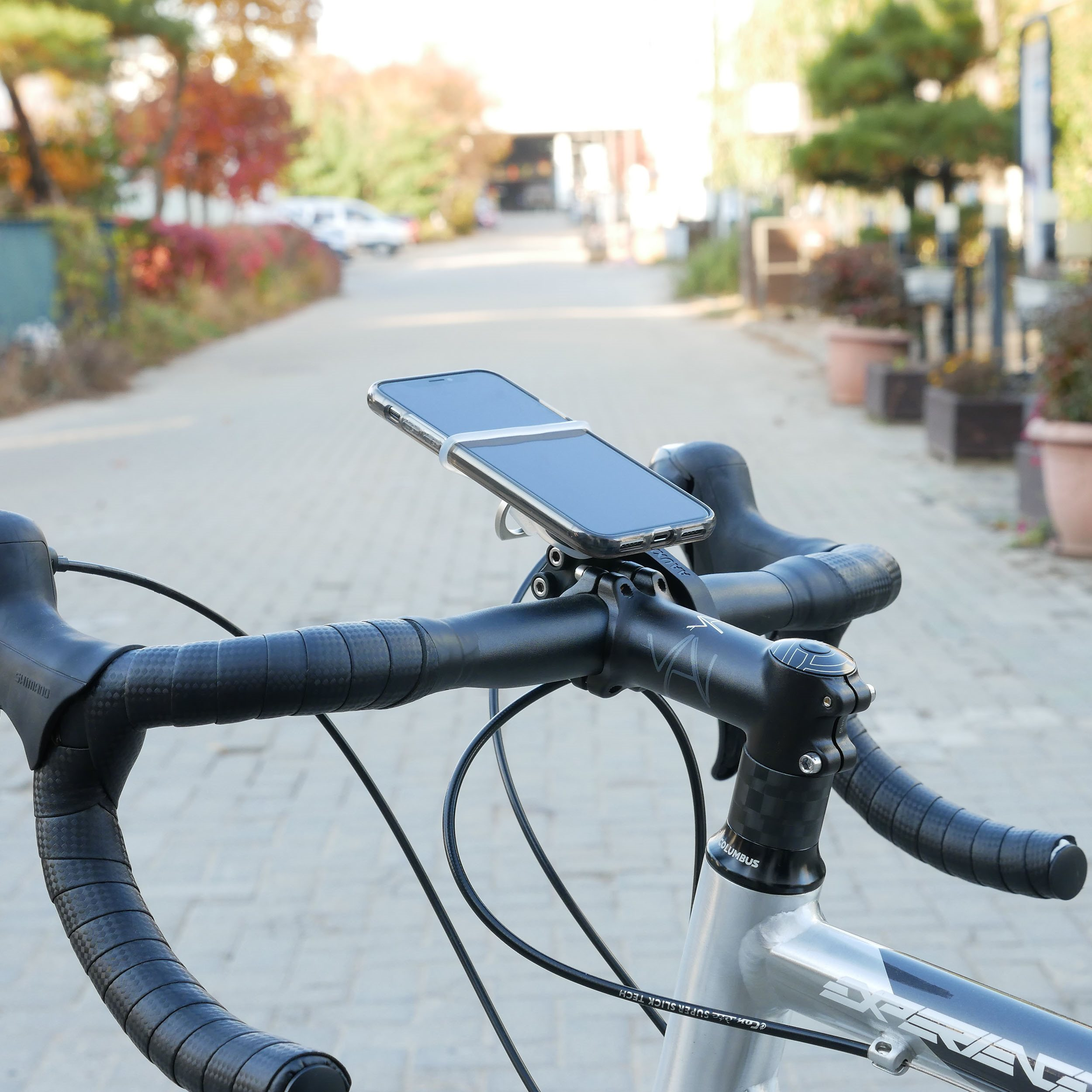 Telefon an Fahrradlenker montiert. Silbernes Fahrrad mit schwarzem Lenker. Hintergrund ist eine gepflasterte Straße und Laub.