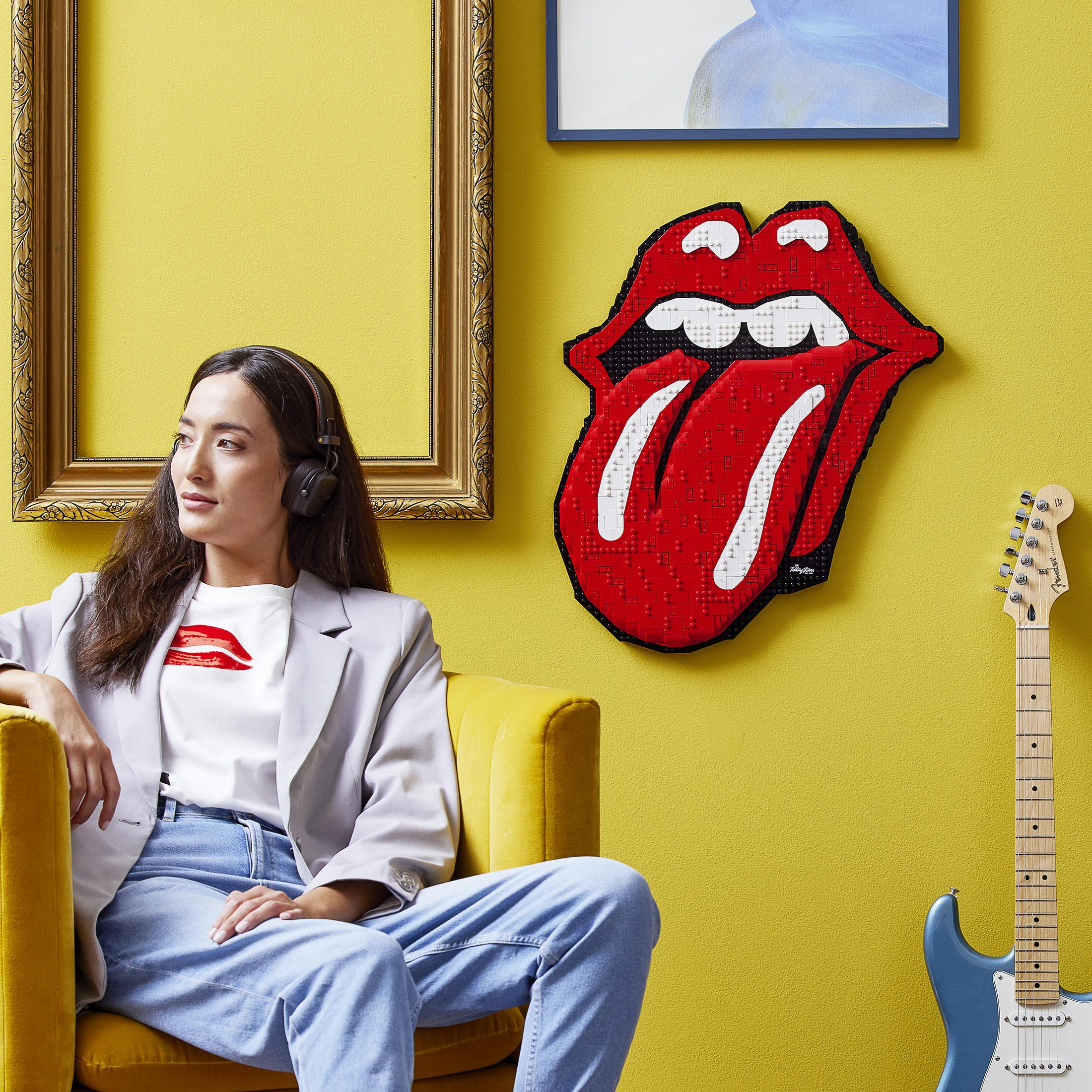 Kobieta w pokoju z żółtą ścianą, w słuchawkach, siedząca obok gitary i logo Rolling Stones.