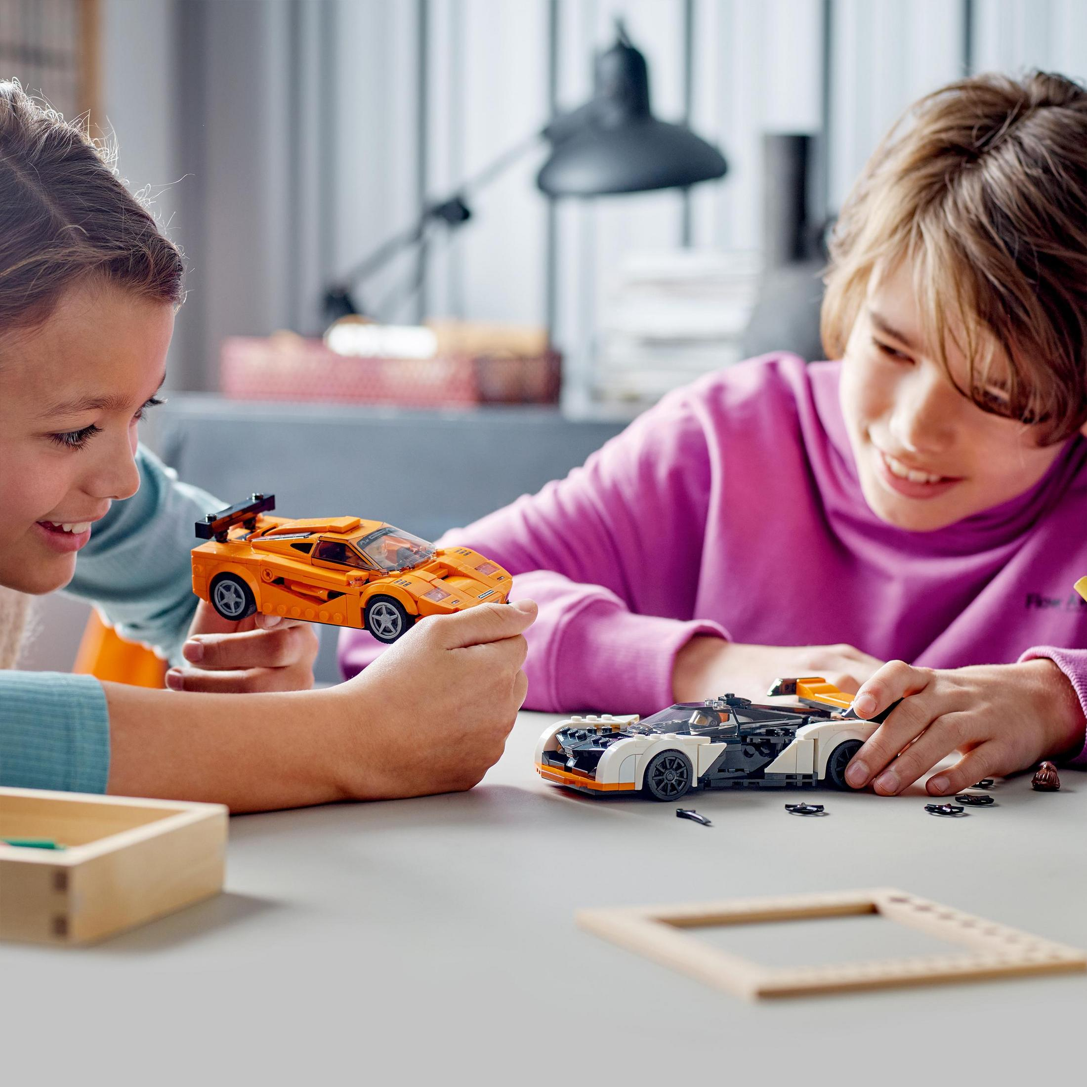Zwei Kinder spielen mit Spielzeugautos auf einem Tisch. Eines hält ein orangefarbenes Auto, das andere ein schwarzes.