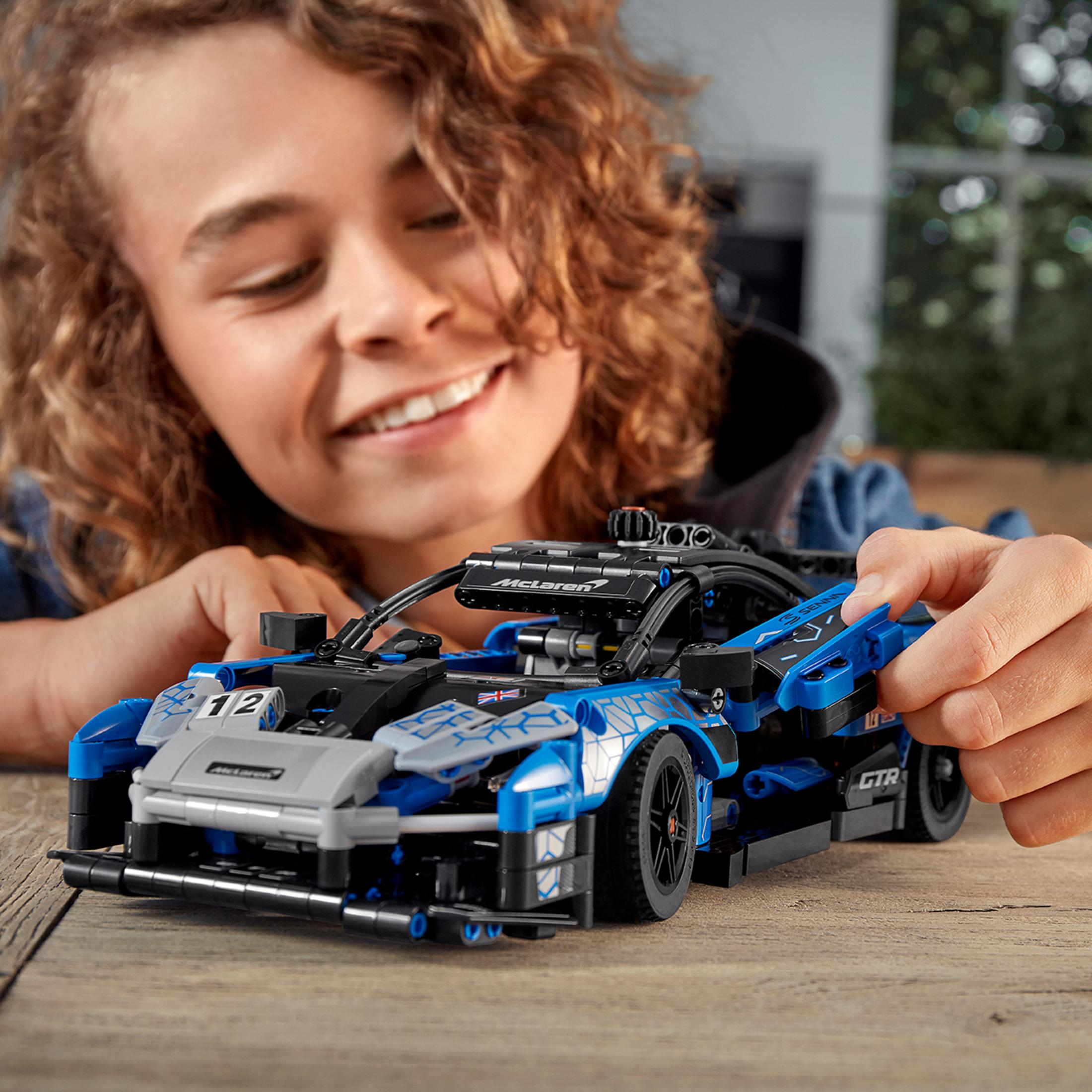 Chłopiec uśmiecha się do niebiesko-czarnego modelu samochodu Lego McLaren. Model stoi na drewnianym stole.