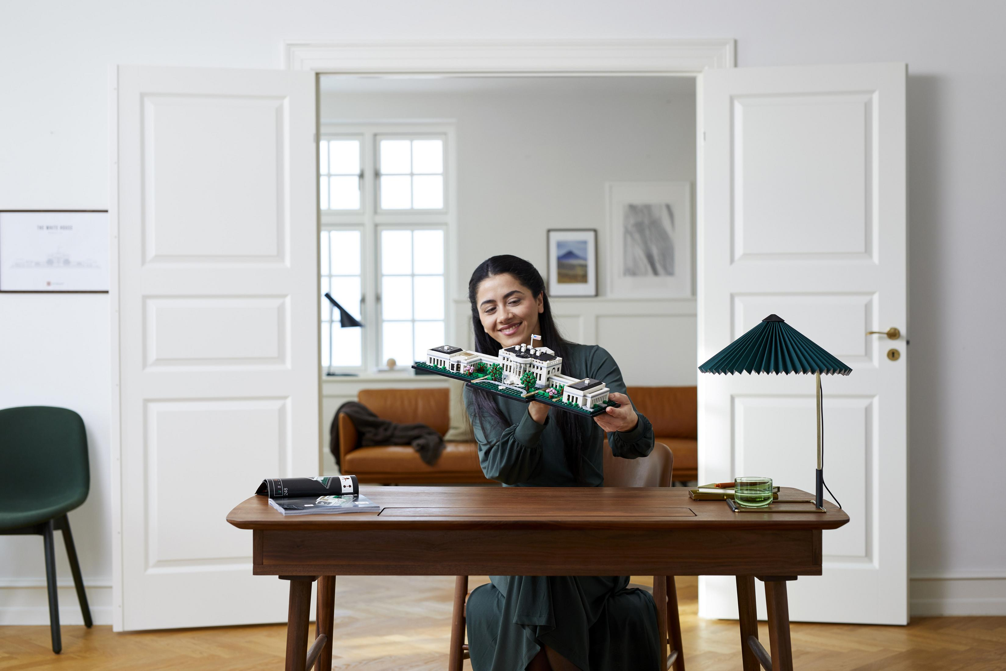 Kobieta trzymająca model budowli Lego. Uśmiecha się. Pokój z otwartymi drzwiami i stołem.