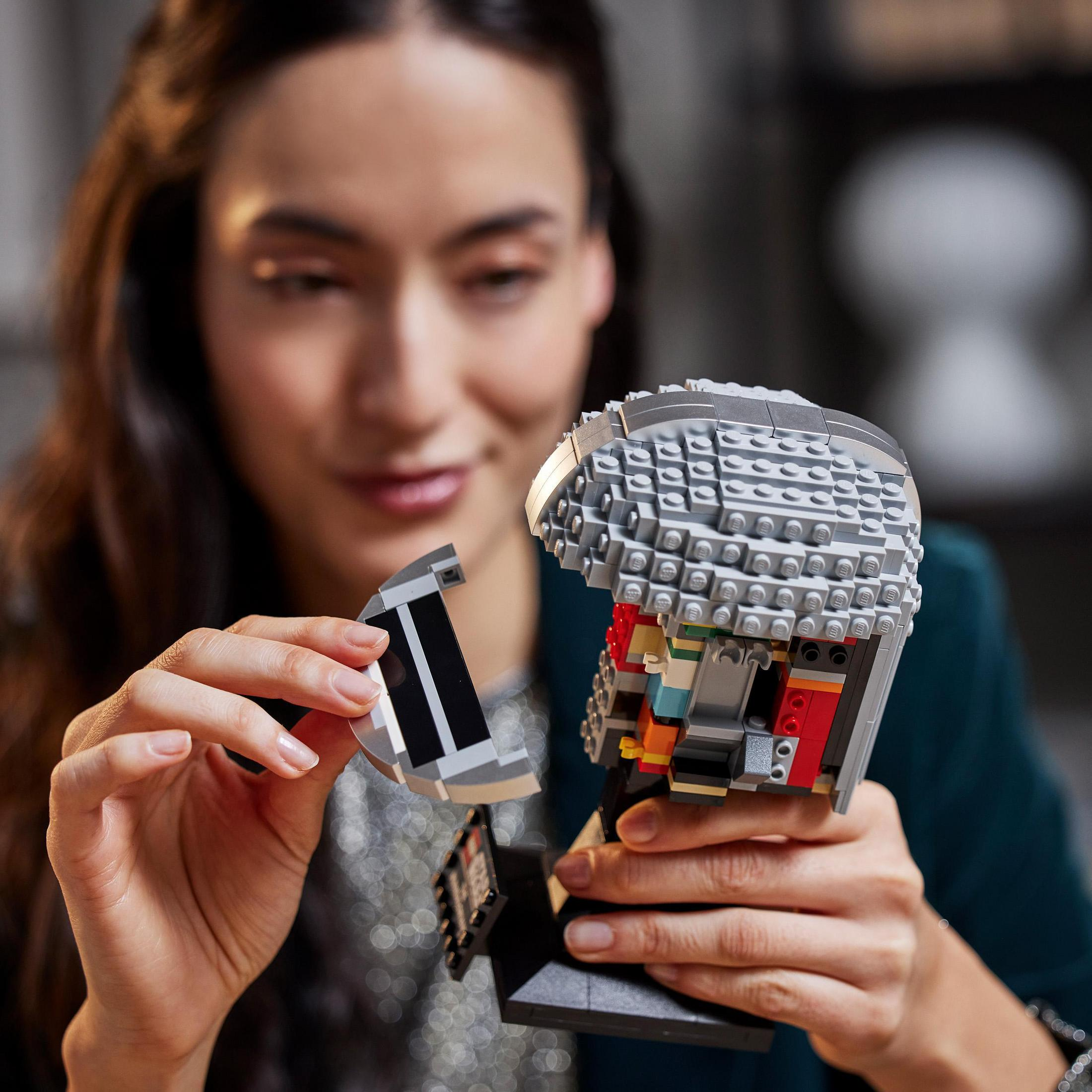 Kobieta buduje hełm Lego, trzymając element. Białe tło.