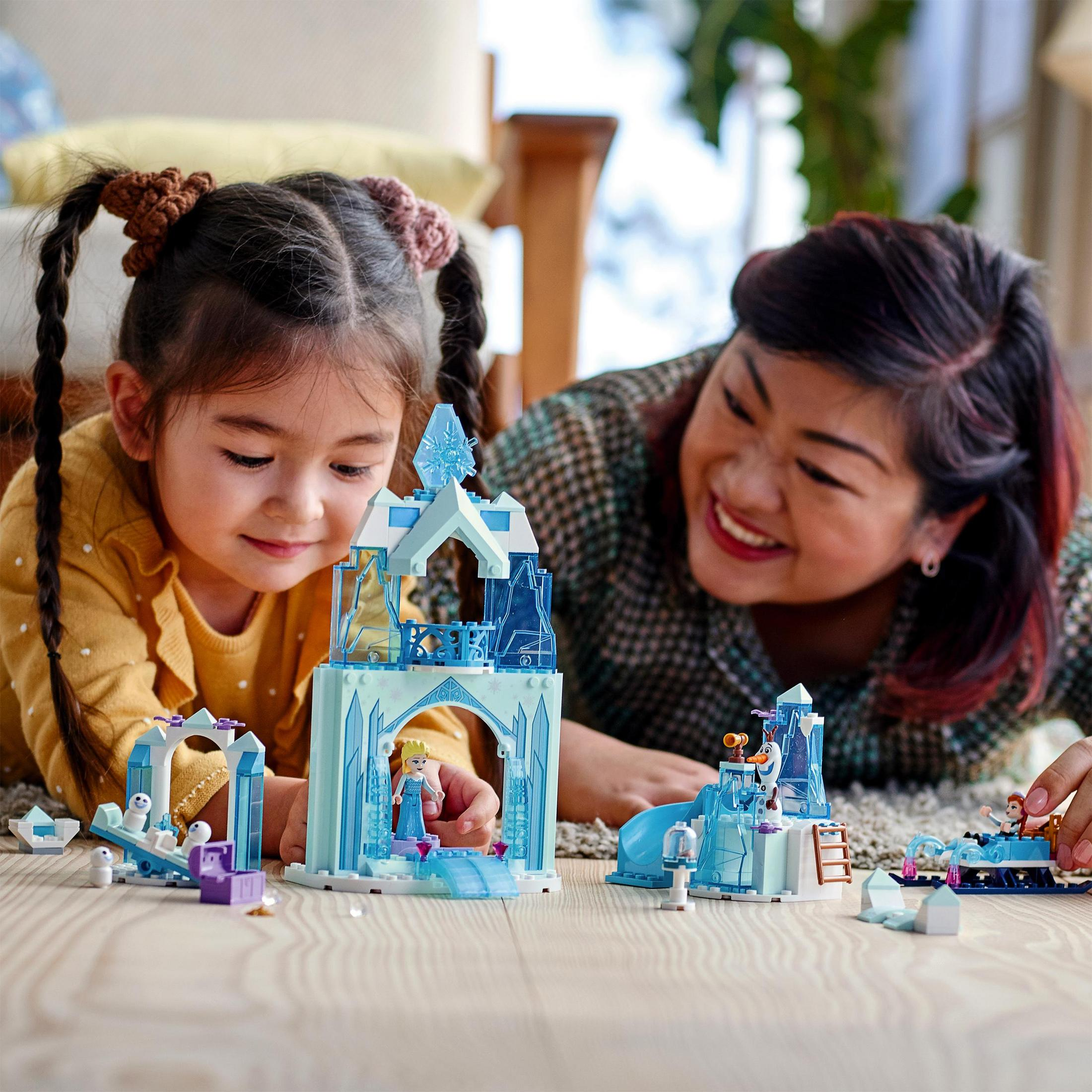 Zwei Mädchen spielen mit einem Lego-Set, mit einem Burgmodell und einer lächelnden Frau im Hintergrund.