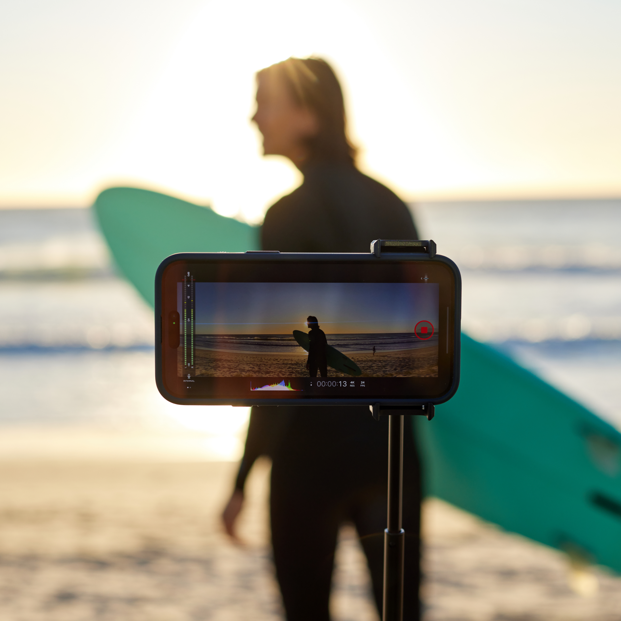 Ein Telefon zeichnet einen Mann mit Surfbrett am Strand auf. Der Bildschirm zeigt eine Videooberfläche. Sonnenuntergang im Hintergrund.