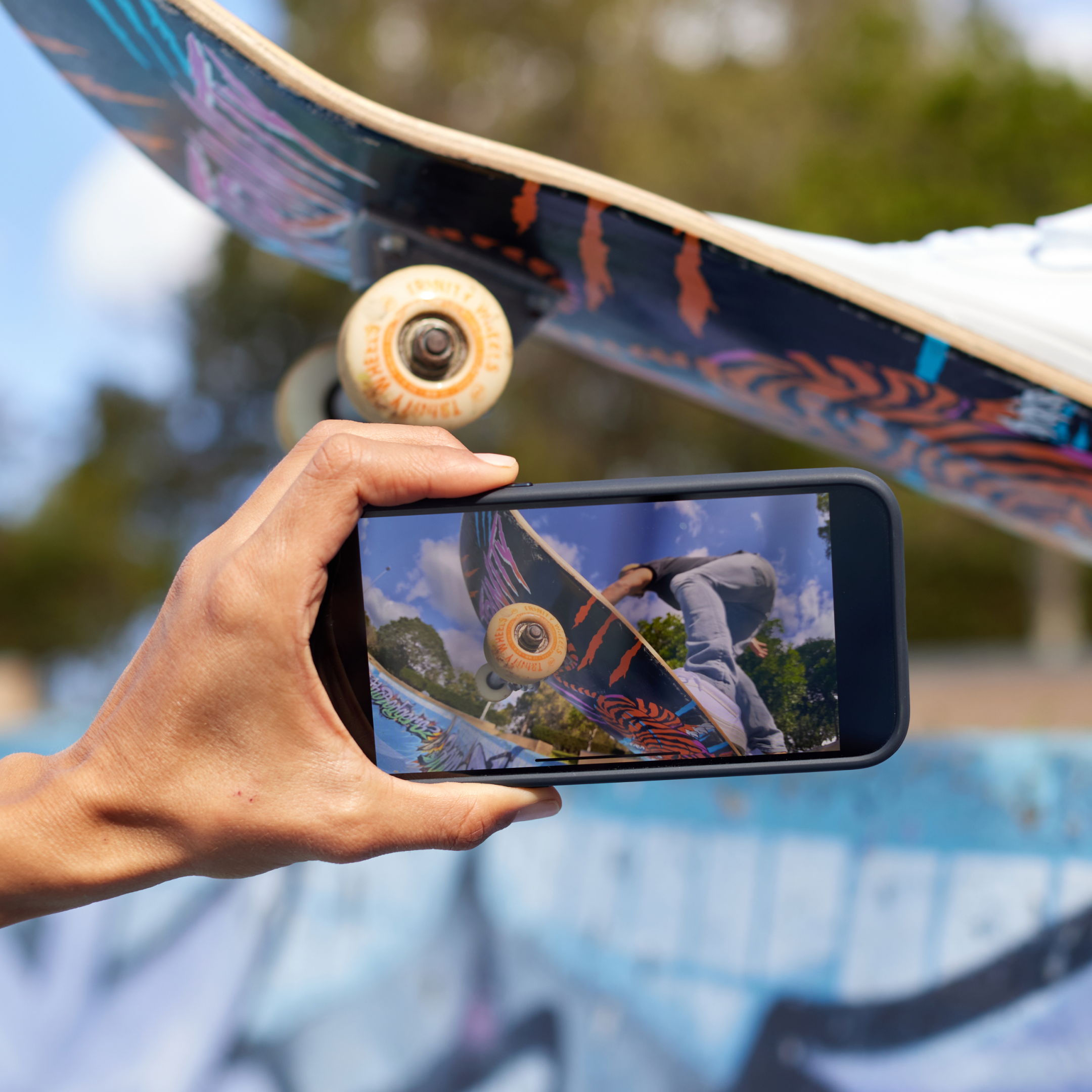 Eine Hand, die ein Telefon hält und einen Skateboarder in der Luft zeigt. Ein echtes Skateboard ist darüber.