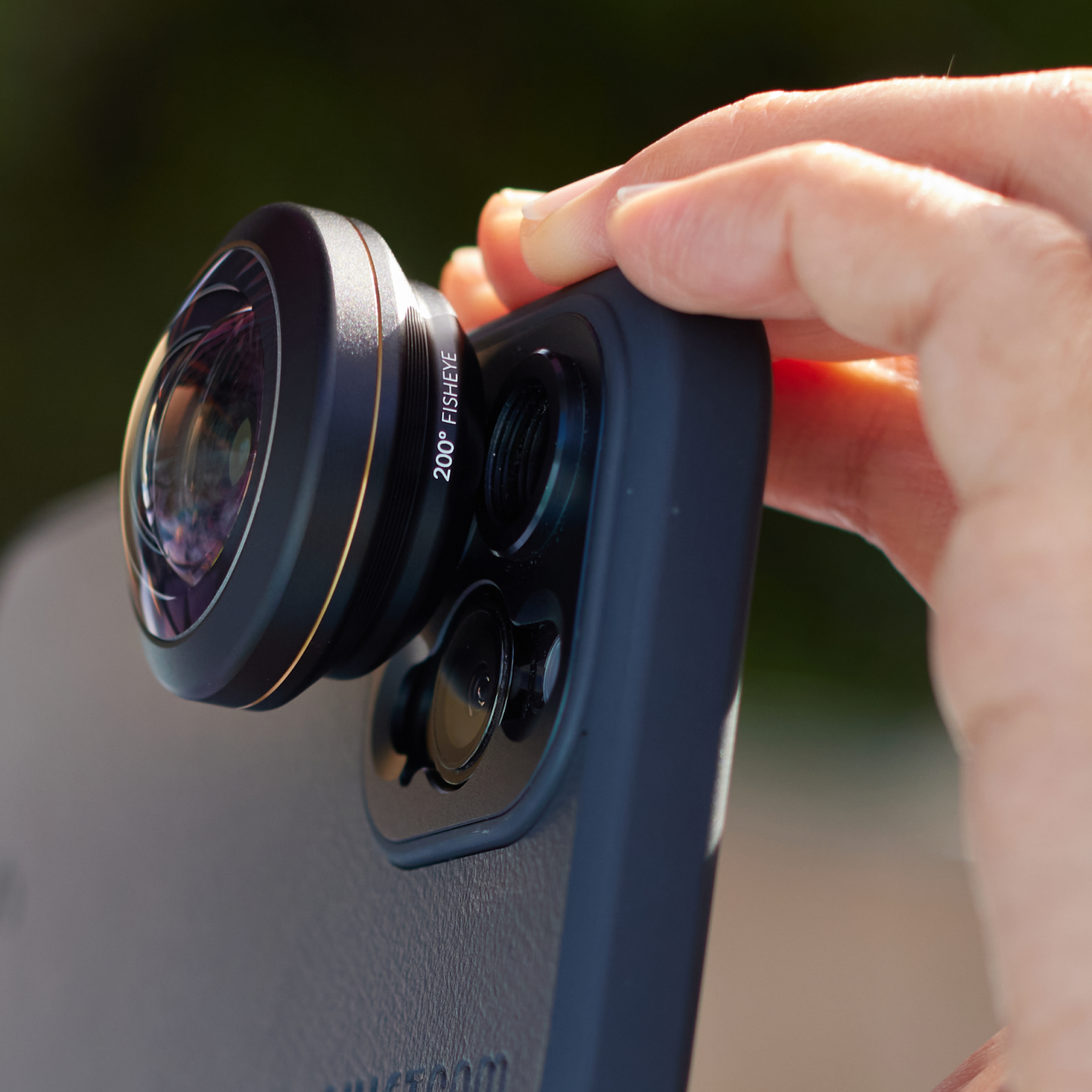 Hand, die ein Telefon mit einem Fisheye-Objektivaufsatz hält, vor einem verschwommenen Hintergrund.