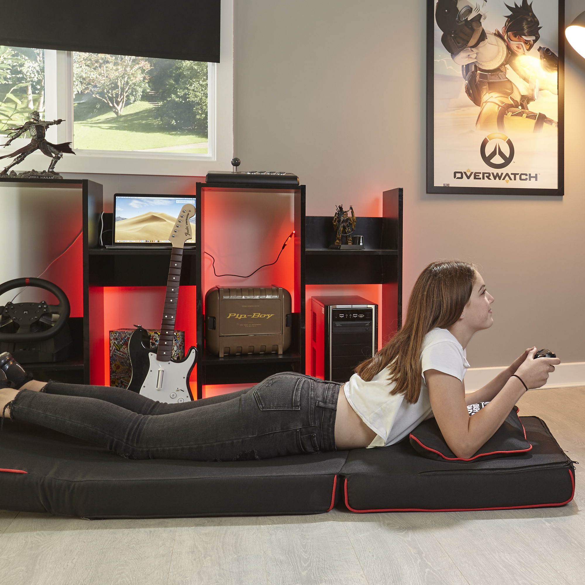 Ein Mädchen liegt auf einer schwarzen Matte und spielt ein Videospiel. Laptop und Gitarre stehen daneben.