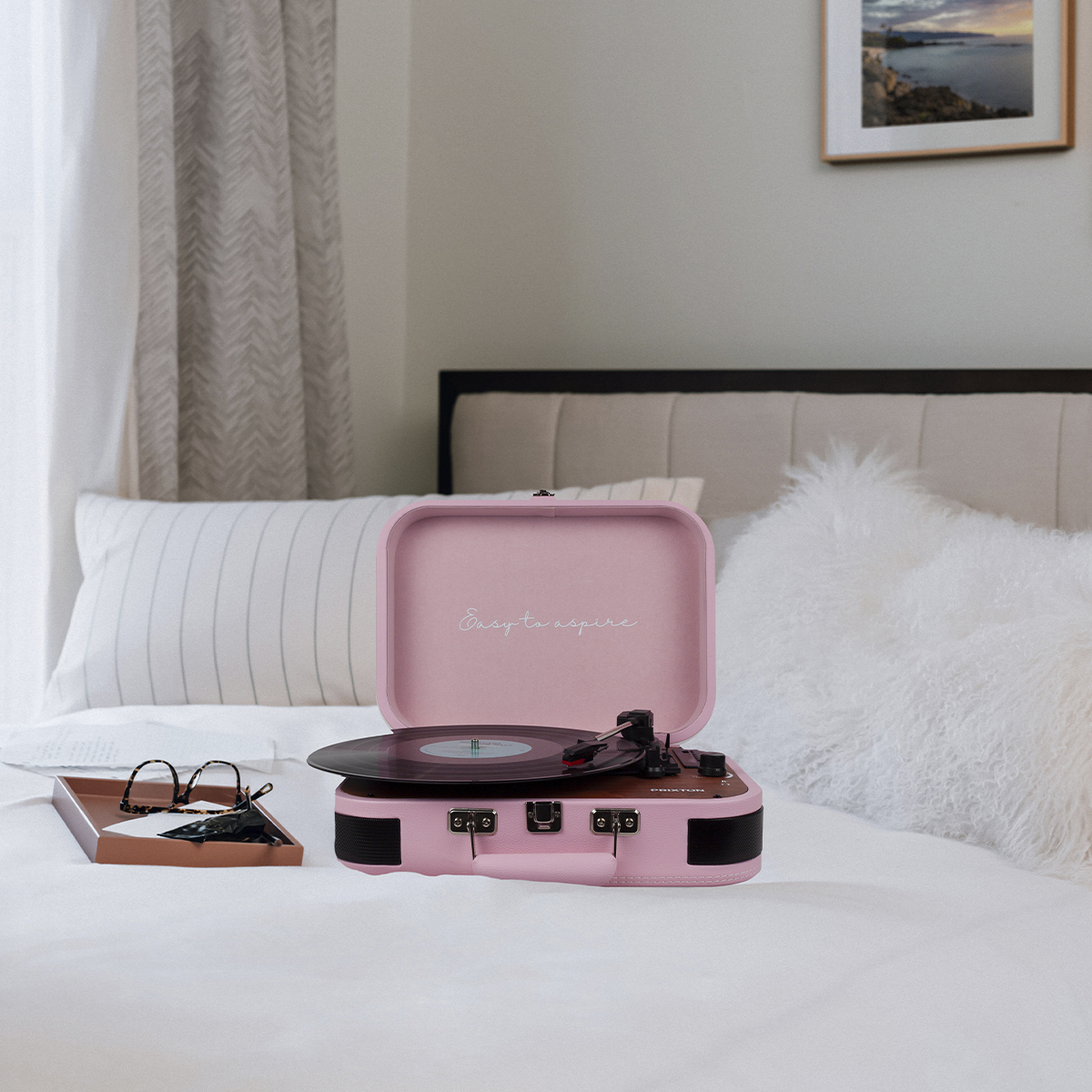 Różowy gramofon na łóżku, otwarty z płytą. Okulary i taca w pobliżu.