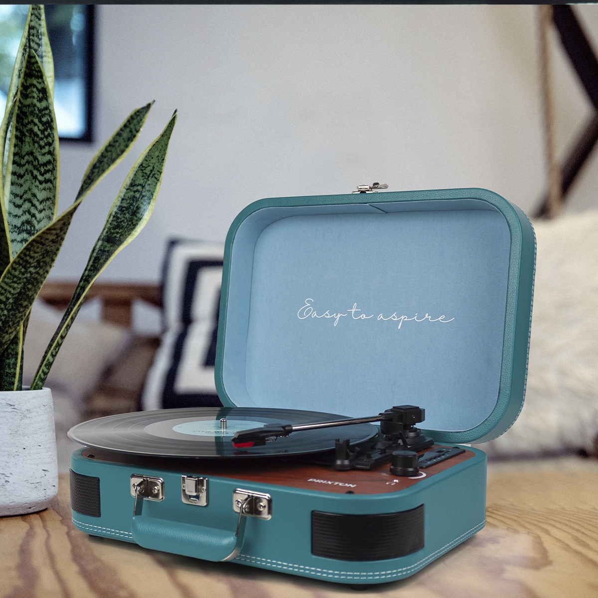 Turkusowy gramofon z otwartą pokrywą i grającą płytą. Drewniane i czarne elementy sterujące. Tekst: Łatwo dążyć.