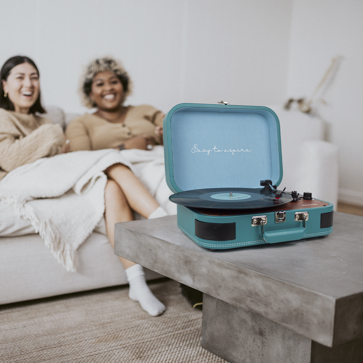 Turkusowy gramofon otwarty na szarym stole. Dwie kobiety w tle.