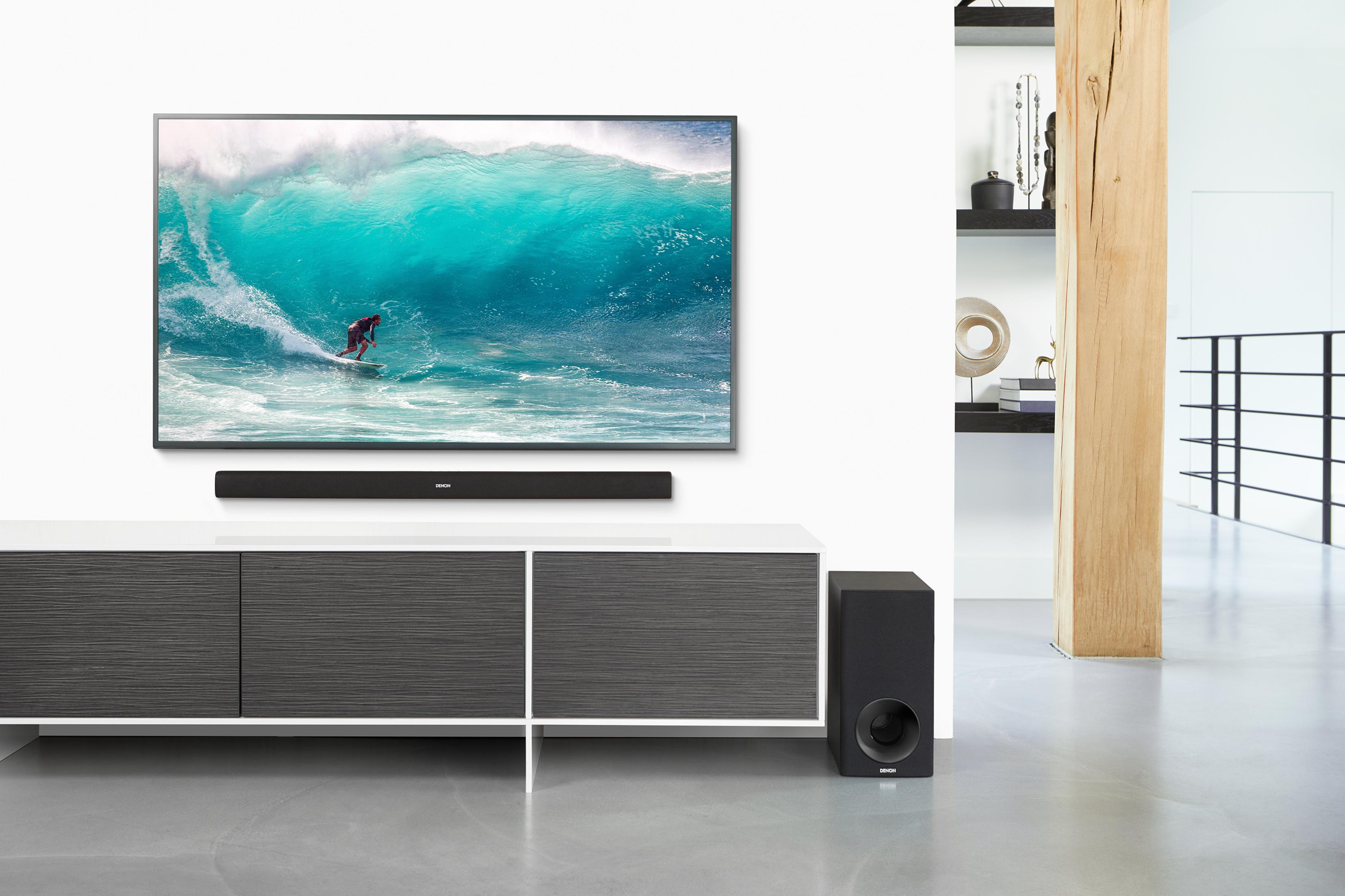 Fernseher mit Surfer auf dem Bildschirm, Soundbar darunter. Weiße Wand, Regal und Subwoofer rechts.
