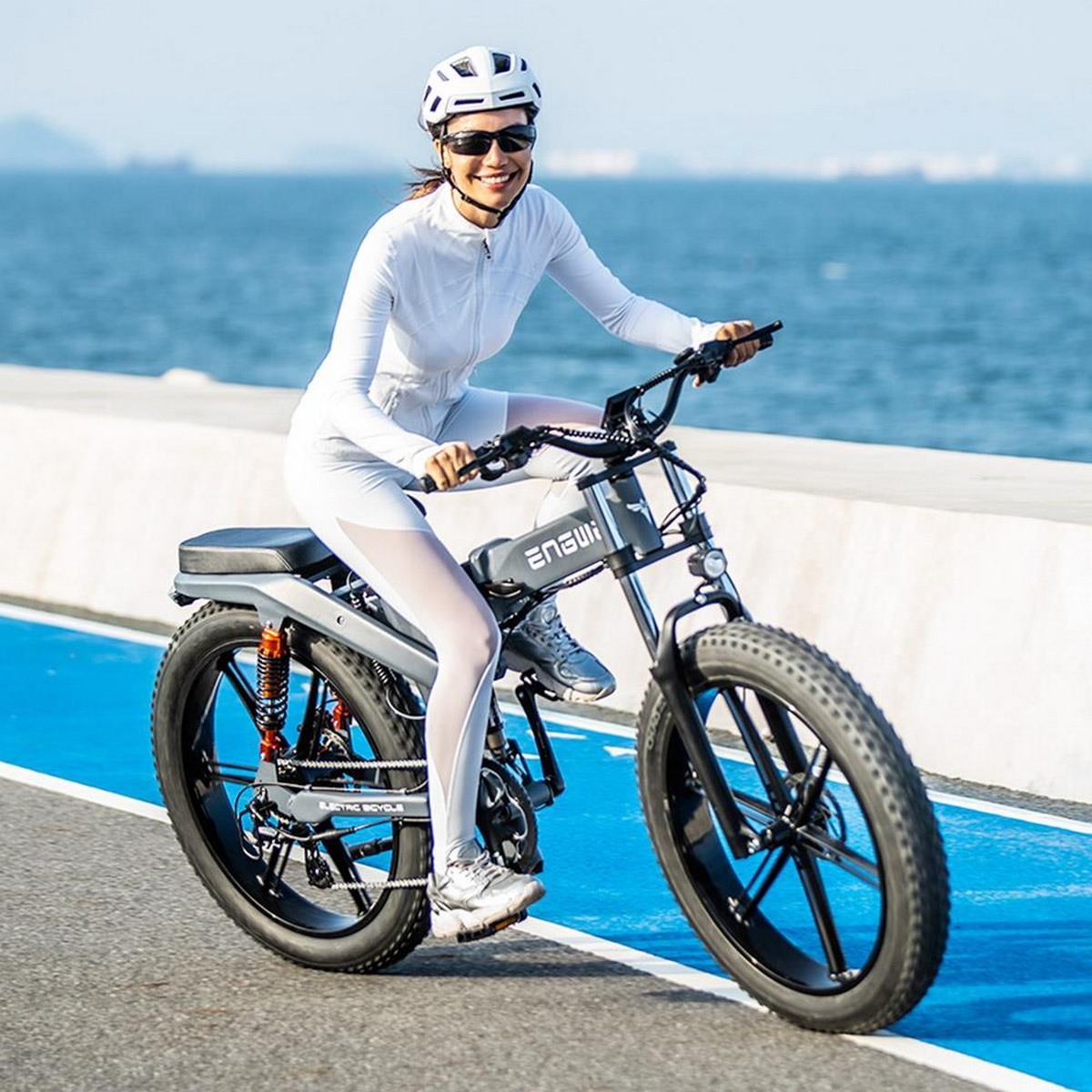 Frau fährt E-Bike auf blauem Weg am Meer, mit Helm und Sonnenbrille.