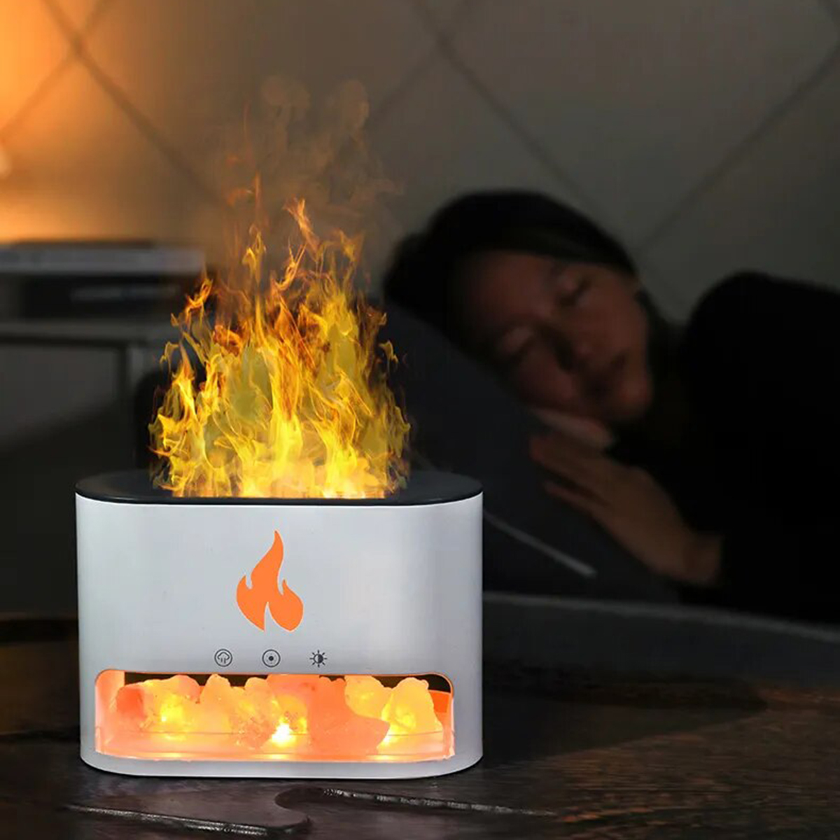 Una lámpara LED blanca con forma de llama y llamas naranjas, junto a una persona durmiendo en la cama.