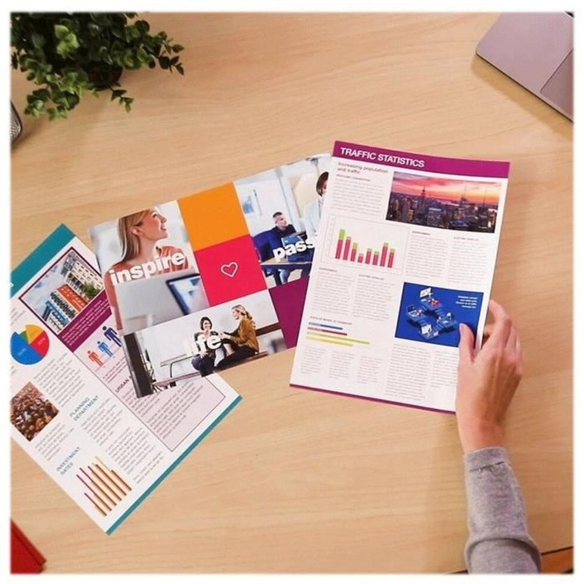 Dokumenty na drewnianym stole: magazyn, broszury i dłoń trzymająca dokument. Tło: roślina i laptop.