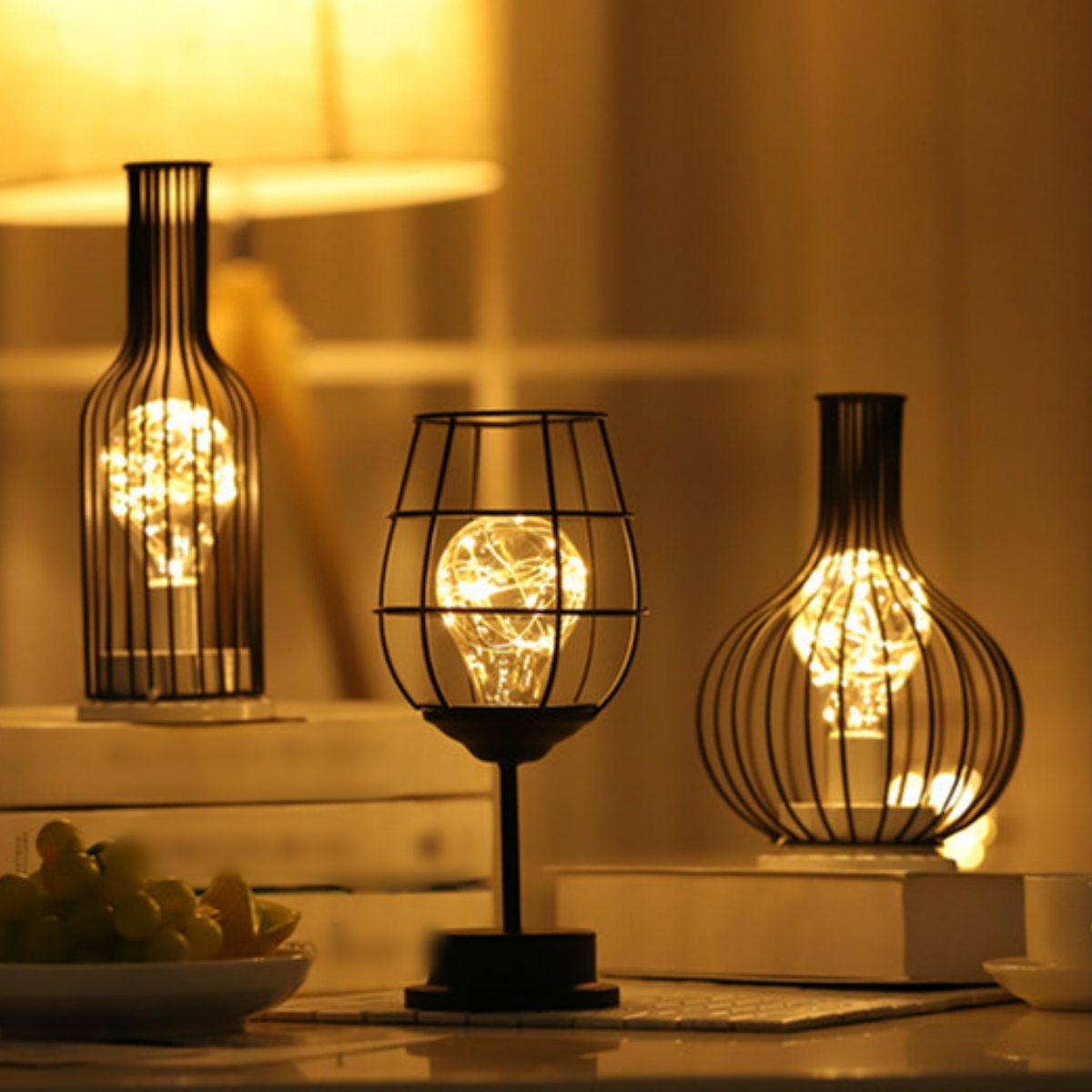 Drei Lampen mit Glühbirne im Käfig. Die Lichter stehen auf einem Tisch mit Büchern und einem Teller.
