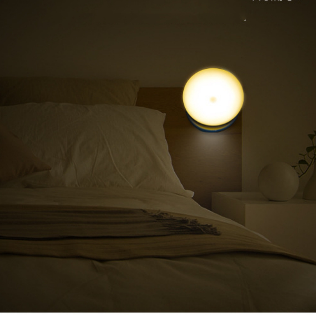 Eine runde, gelbe Leuchte an einer Wand über einem Bett. Ein weißes Kopfteil und ein Nachttisch sind sichtbar.