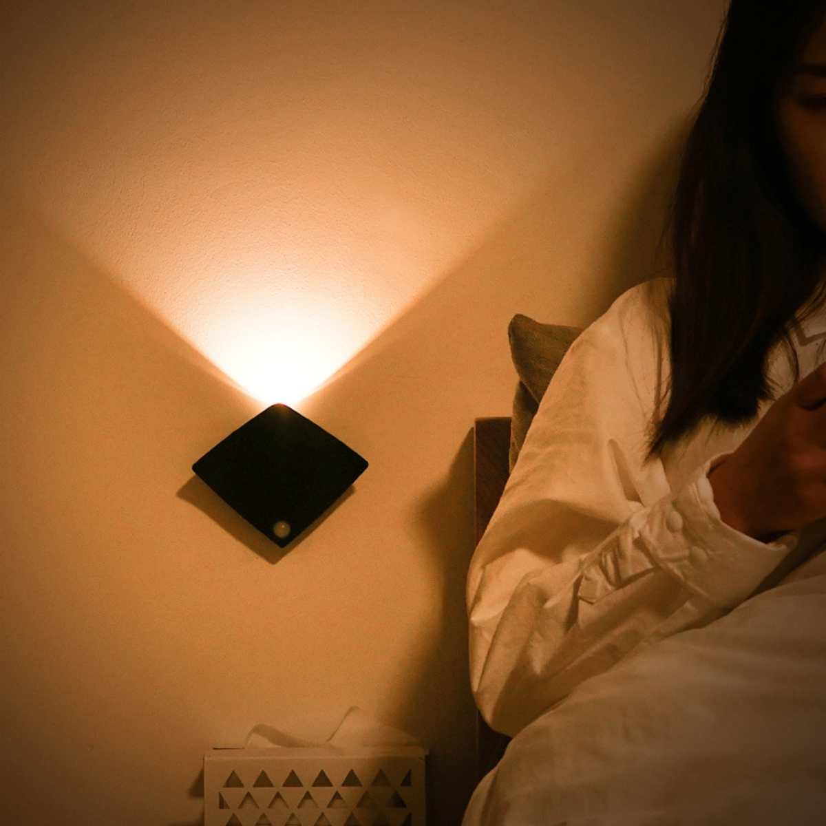 Eine Wandleuchte beleuchtet ein Schlafzimmer. Eine Person sitzt auf dem Bett und benutzt ein Telefon.