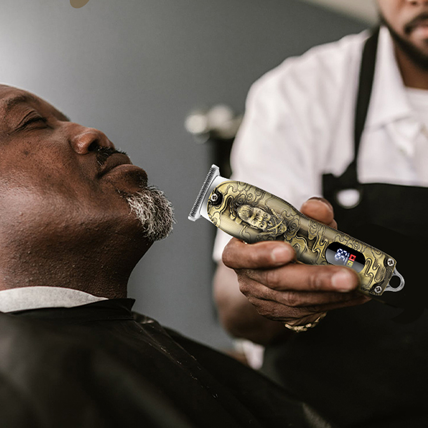 Friseur verwendet Trimmer am grauen Bart eines Mannes. Der Trimmer hat ein Golddesign und Digitalanzeige.