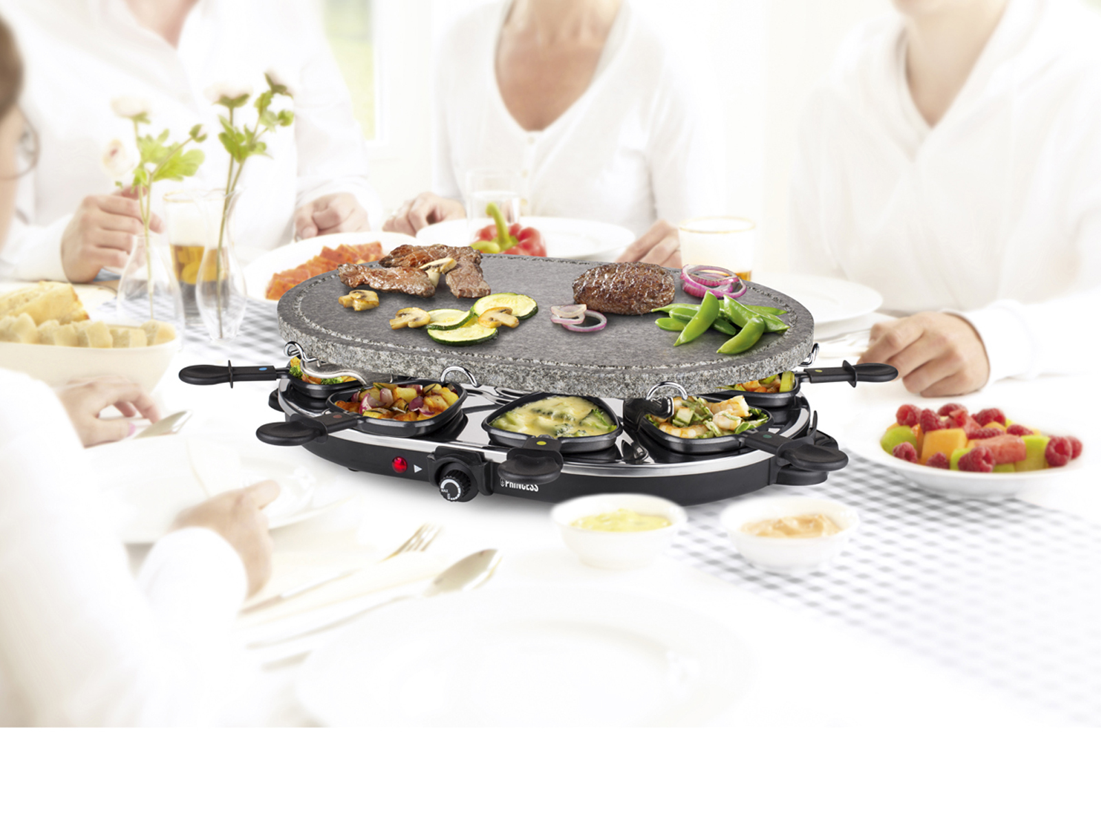 Leute um einen Tisch mit einem Raclette-Grill, Essen und Geschirr.