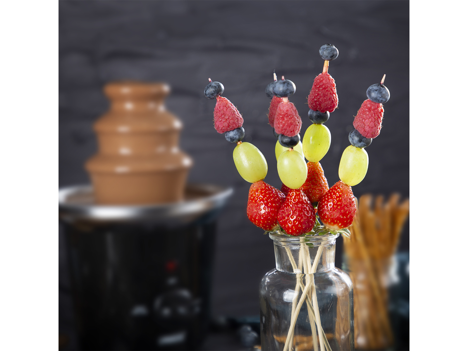 Fruchtspieße, Erdbeeren, Trauben, Himbeeren, Blaubeeren mit Schokoladenbrunnen im Hintergrund.