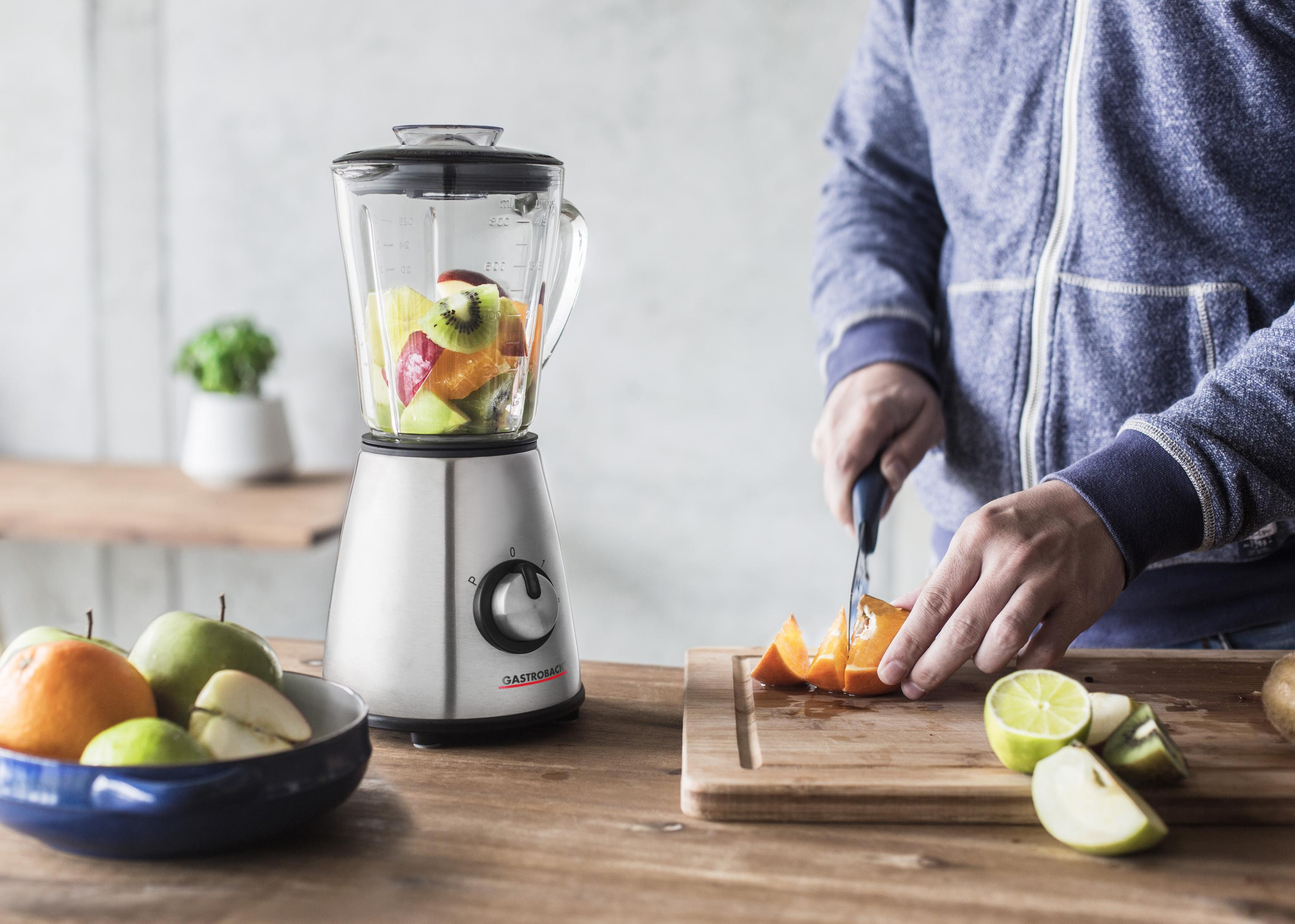 Osoba krojąca pomarańczę, ze blenderem pełnym owoców na drewnianym stole.