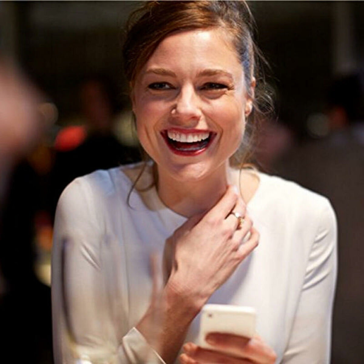 Donna sorridente con telefono. Pelle chiara, capelli castani, camicia bianca. Sfondo sfocato.
