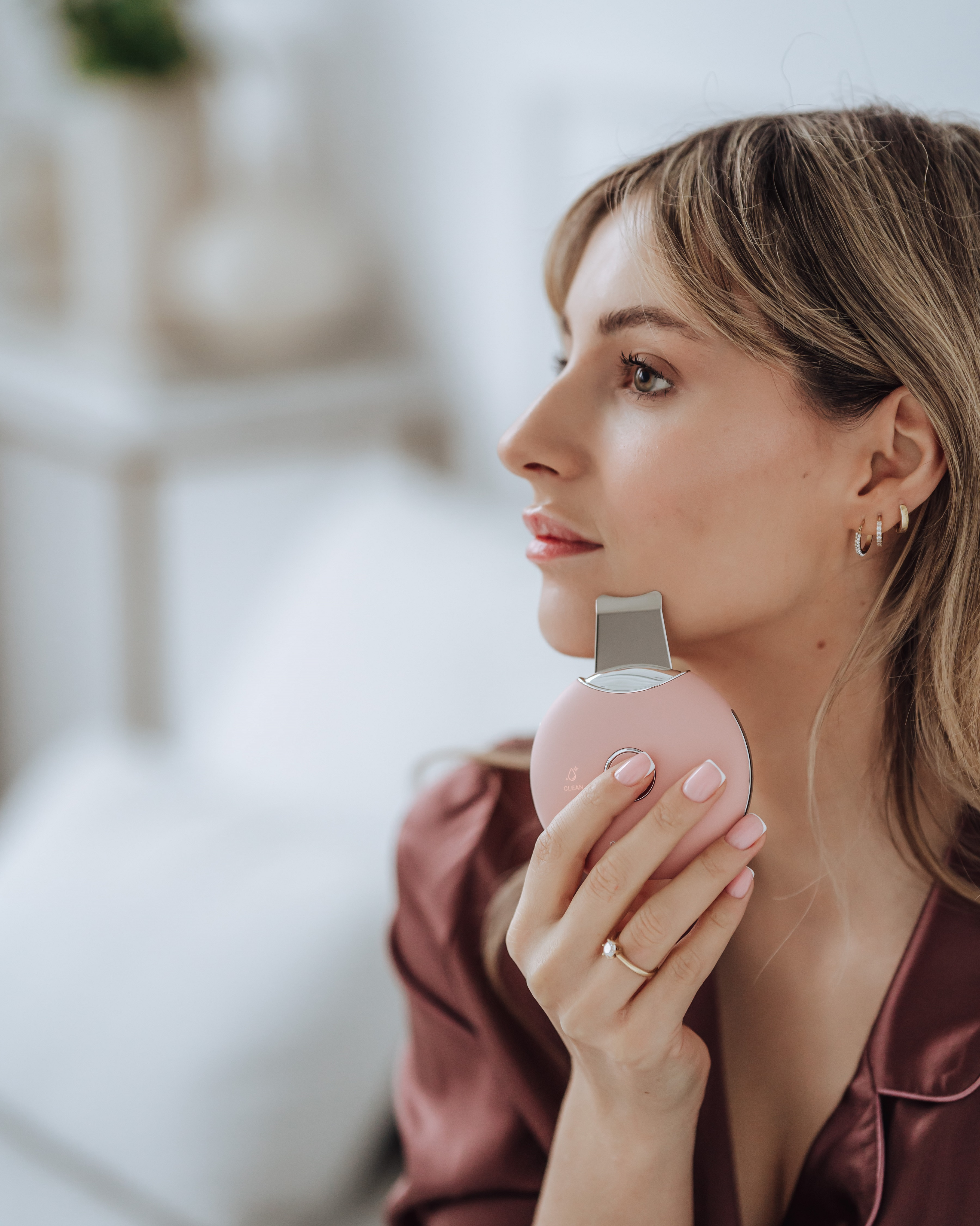 Eine Frau hält ein rosa Gesichtsreinigungsgerät. Sie trägt ein weinrotes Hemd und hat blondes Haar.