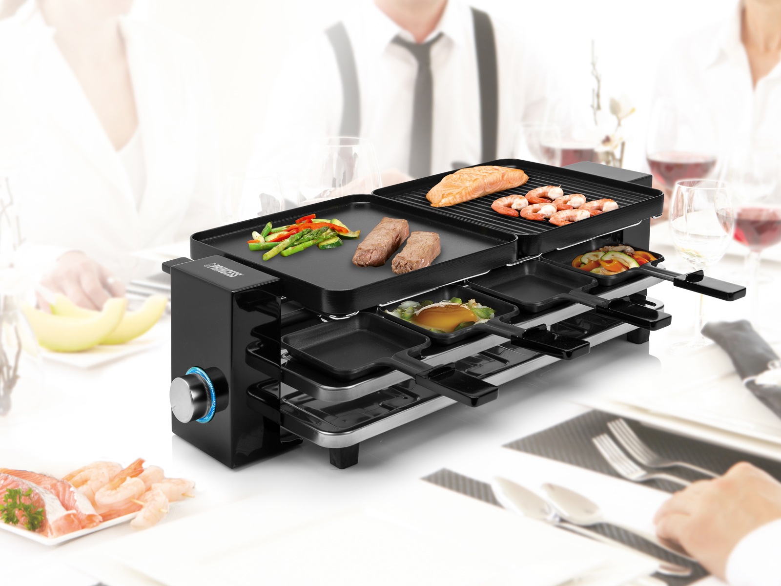 Raclette-Grill mit Essen und Personen im Hintergrund. Der Grill hat verschiedene Gerichte darauf.