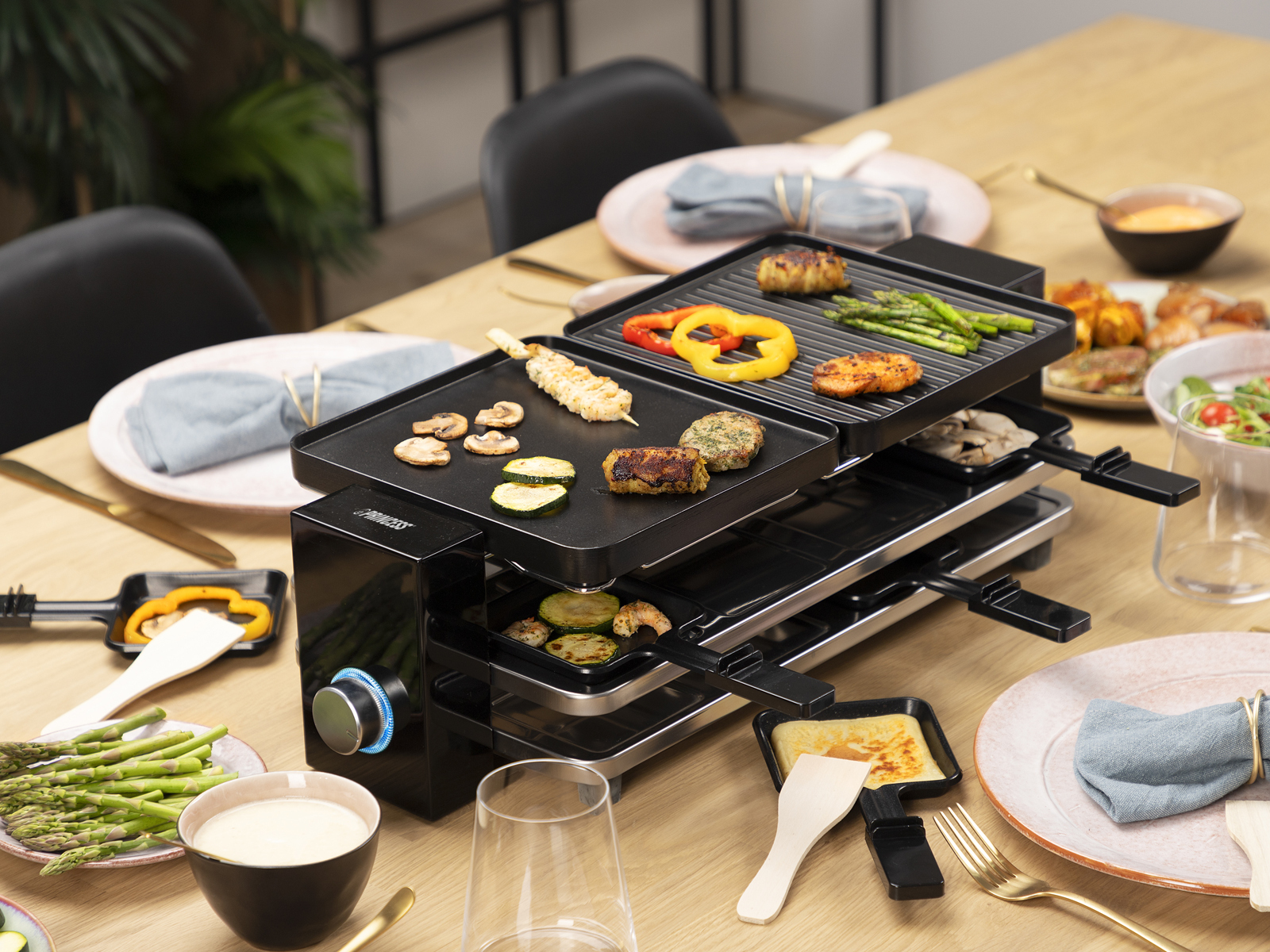 Ein Raclette-Grill mit Essen darauf. Gemüse, Fleisch und Käse sind sichtbar. Tischgedeck ist komplett.