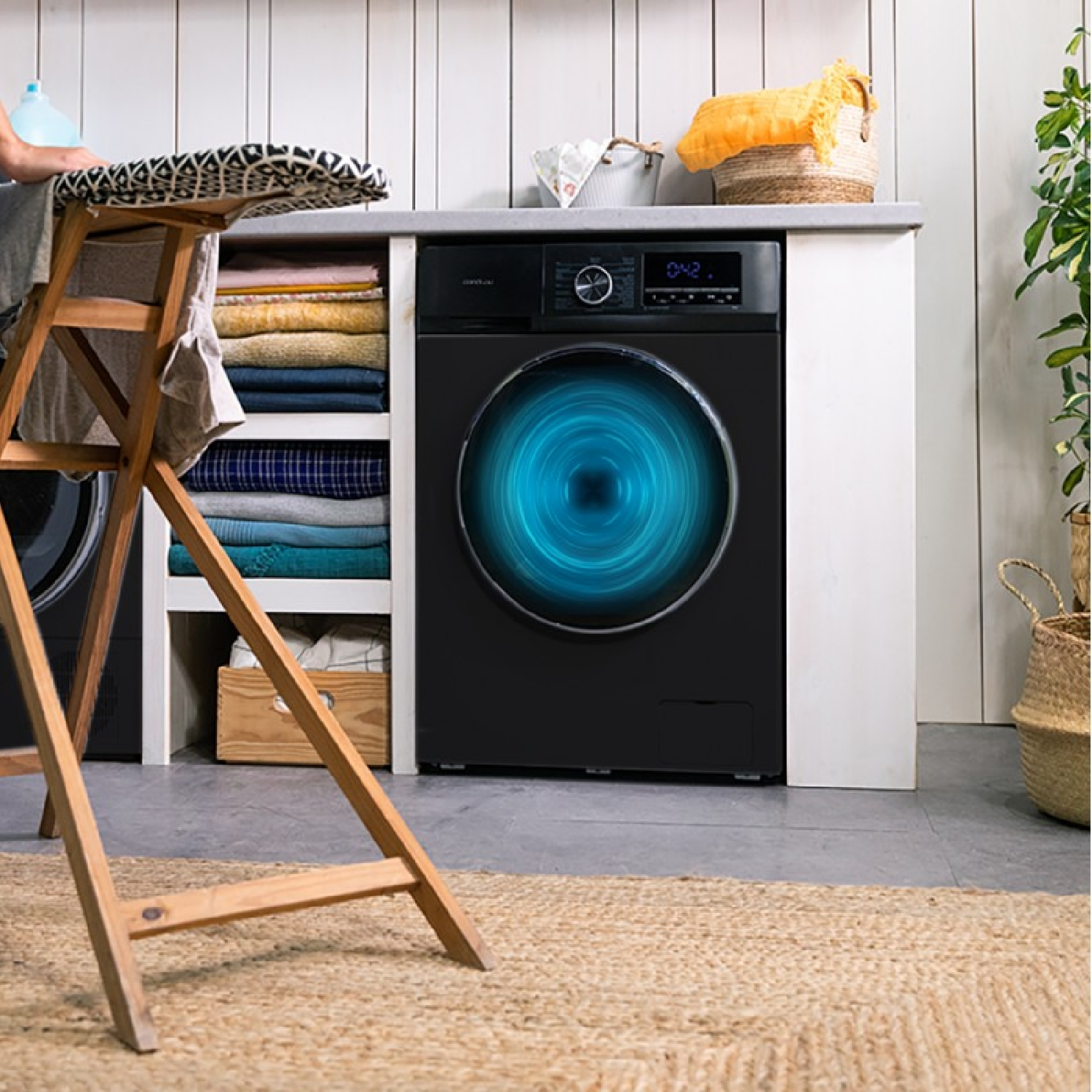 Czarna pralka ze świecącym niebieskim okręgiem. Widoczna deska do prasowania i ubrania.