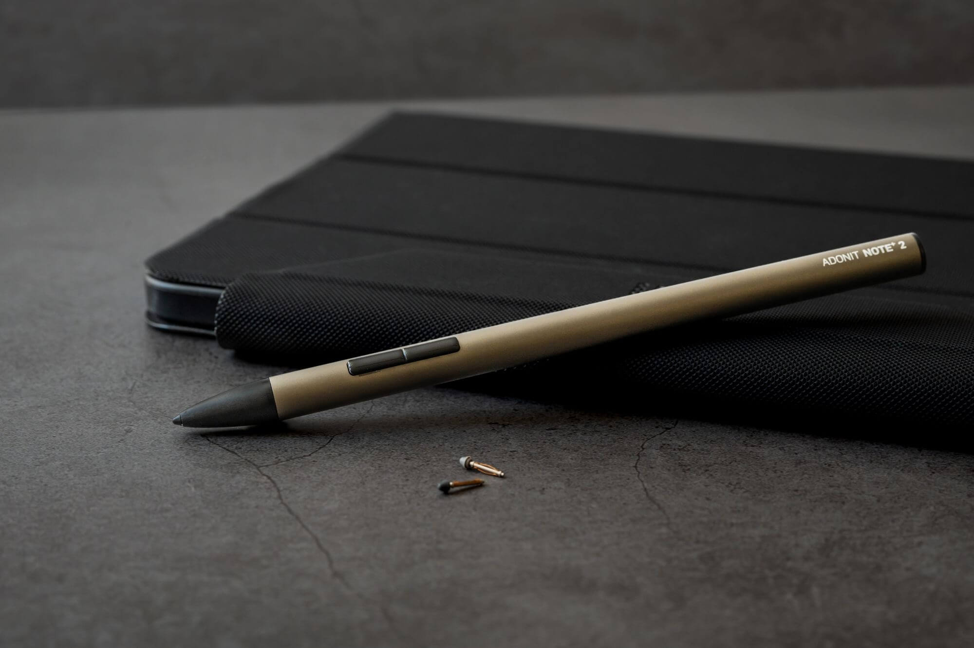 Una penna, etichettata "ADONIT NOTE 2", e due piccole punte su una superficie scura, vicino a un astuccio.