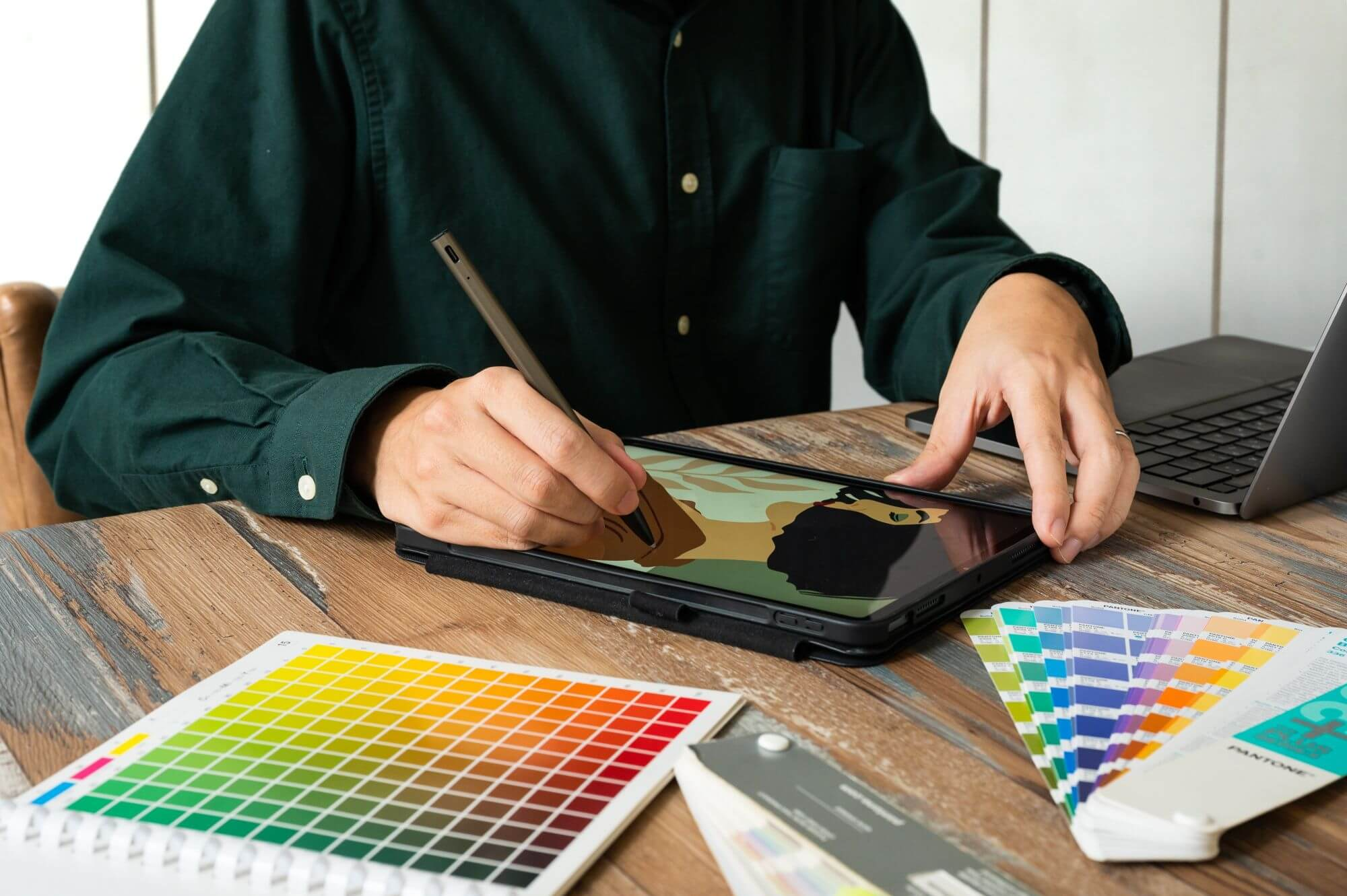 Una persona sta disegnando su un tablet con una penna. Campioni di palette colori e un laptop sono sul tavolo di legno.