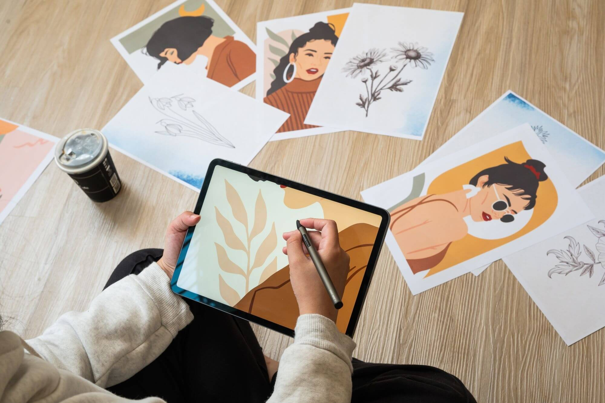 Una persona disegna su un tablet digitale circondata da stampe artistiche e una lattina di caffè.