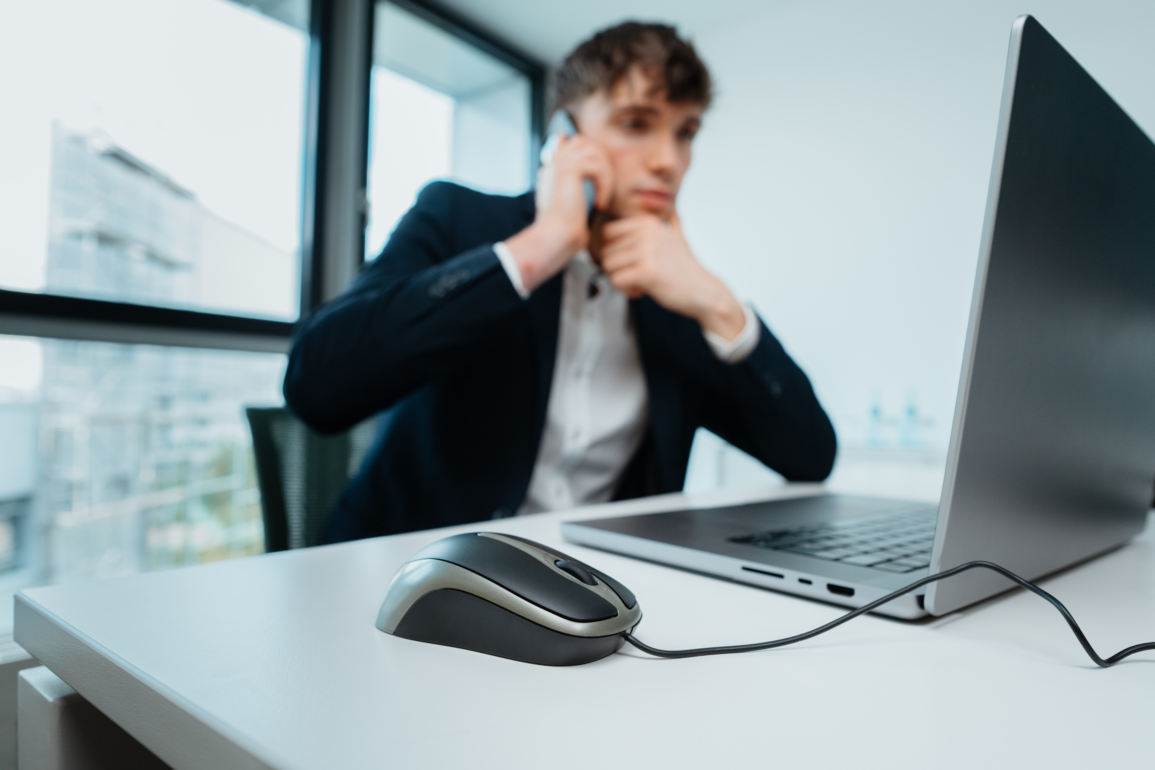 Mężczyzna w garniturze rozmawia przez telefon. Myszka komputerowa leży na stole na pierwszym planie.