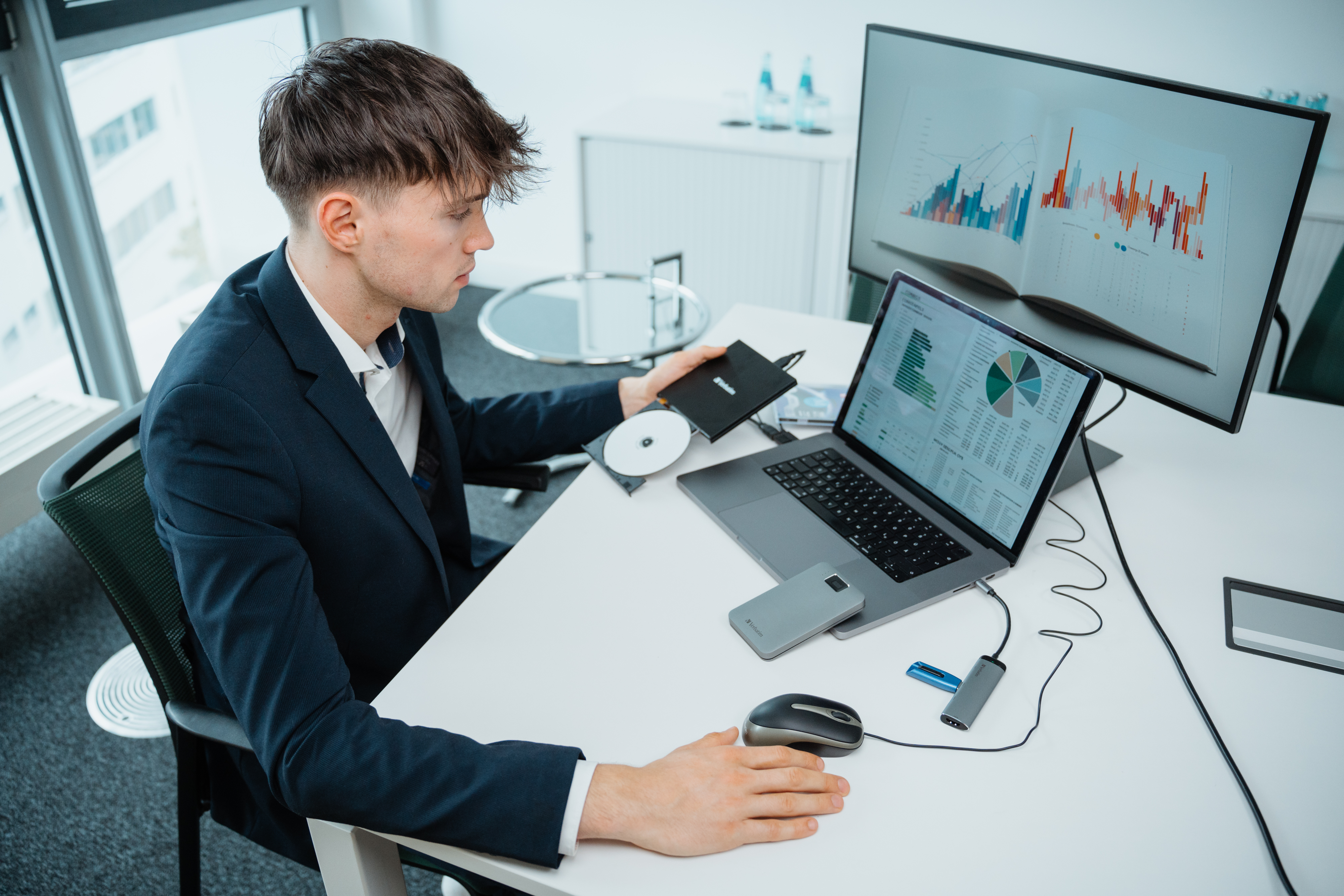 Mężczyzna pracujący przy biurku z laptopem, monitorem i urządzeniami do danych.