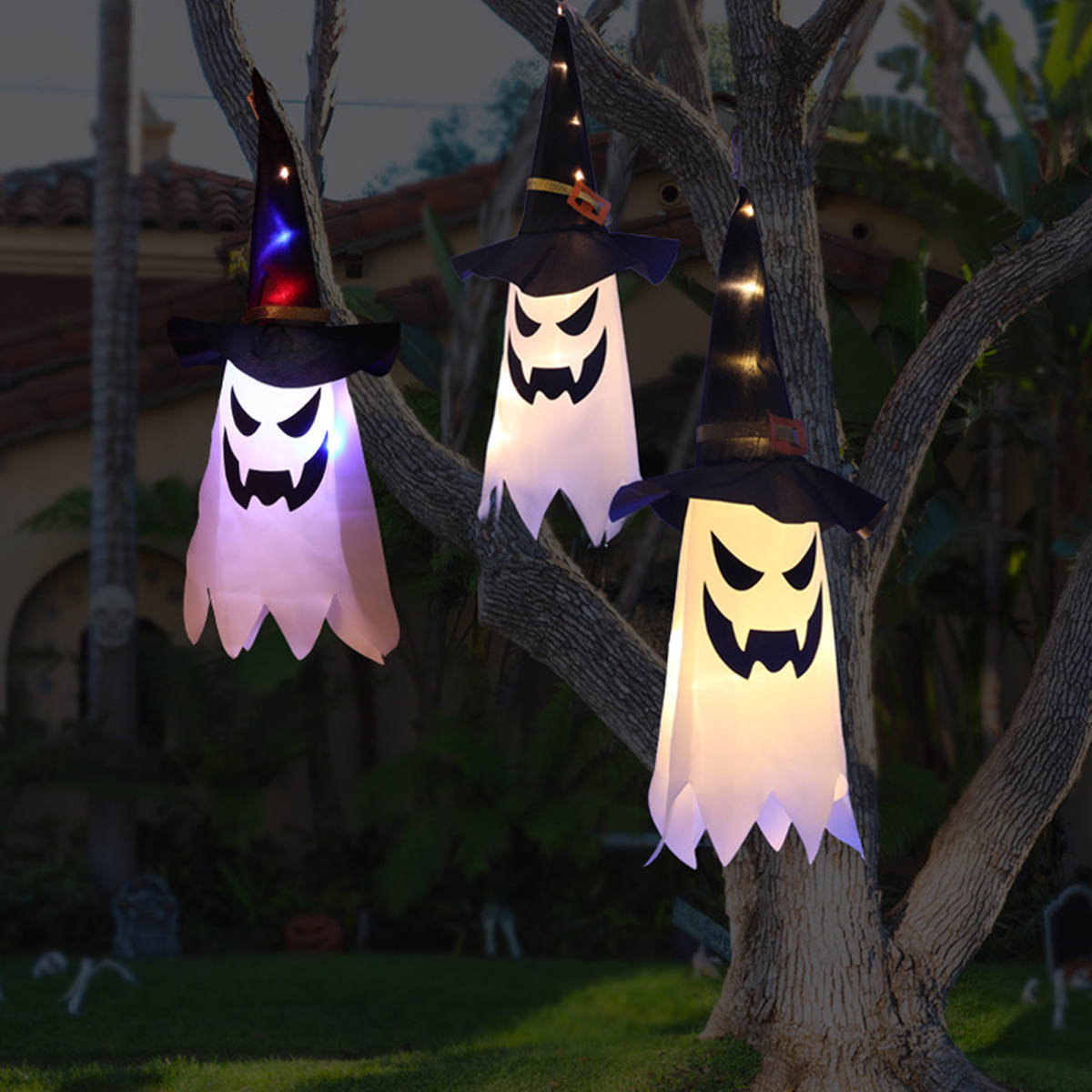 Drei Halloween-Geister-Dekorationen, die an einem Baum hängen. Sie haben Hexenhüte und leuchtende Gesichter.