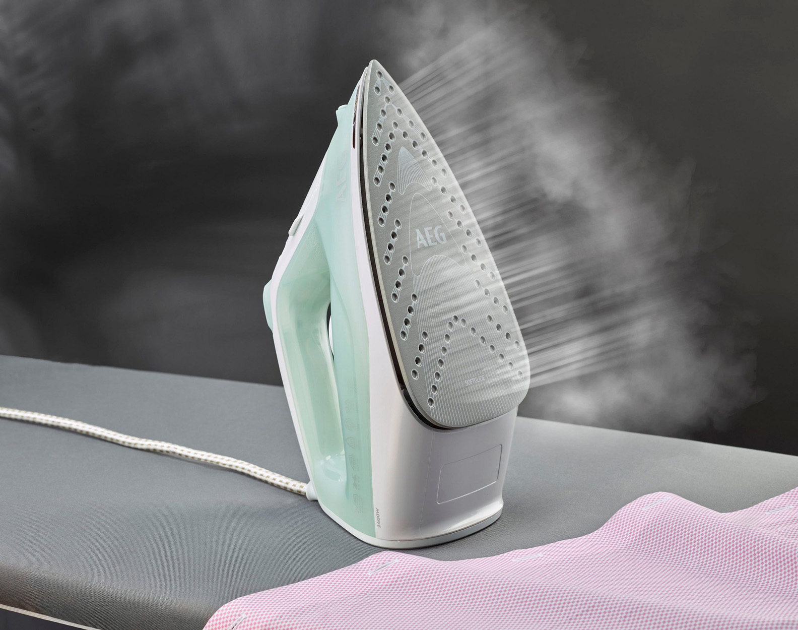 Żelazko parujące czerwoną koszulę na desce do prasowania. Żelazko jest miętowo-zielone i białe.