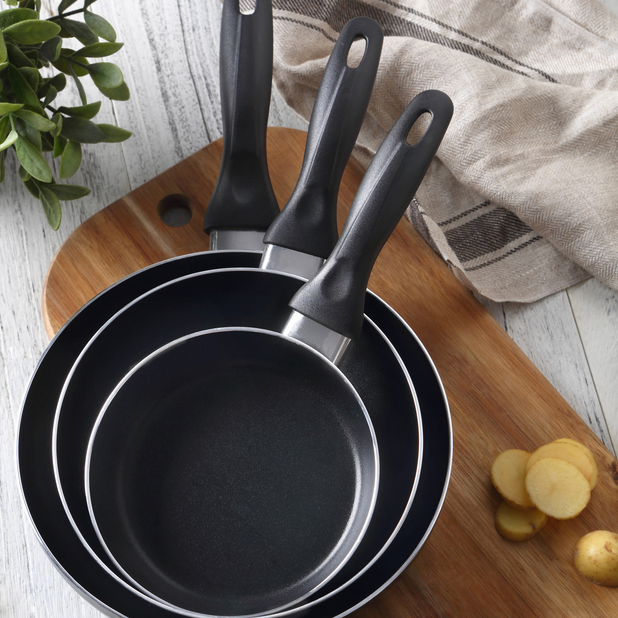 Tres sartenes negras sobre una tabla de madera, con patatas en rodajas y verdura.