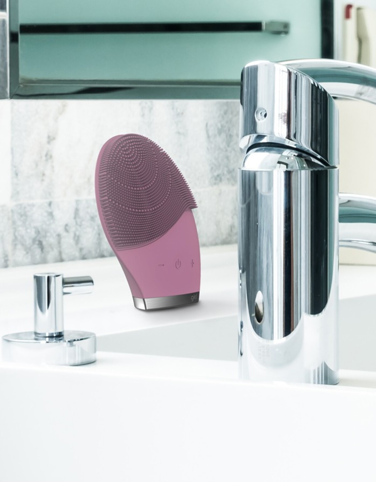 Un dispositivo de limpieza facial rosa en un entorno de baño con un grifo y un lavabo en el fondo.