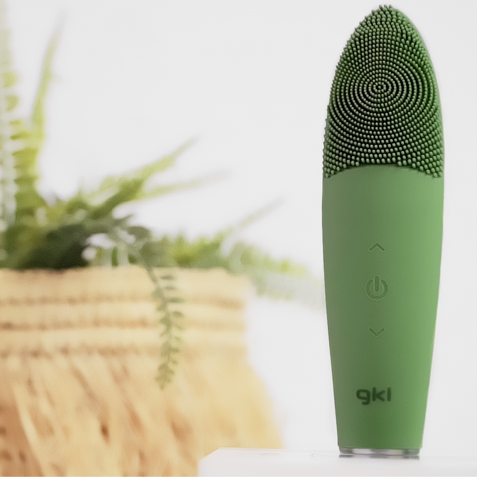 Cepillo de limpieza facial verde con cabezal texturizado. Una planta está en el fondo.