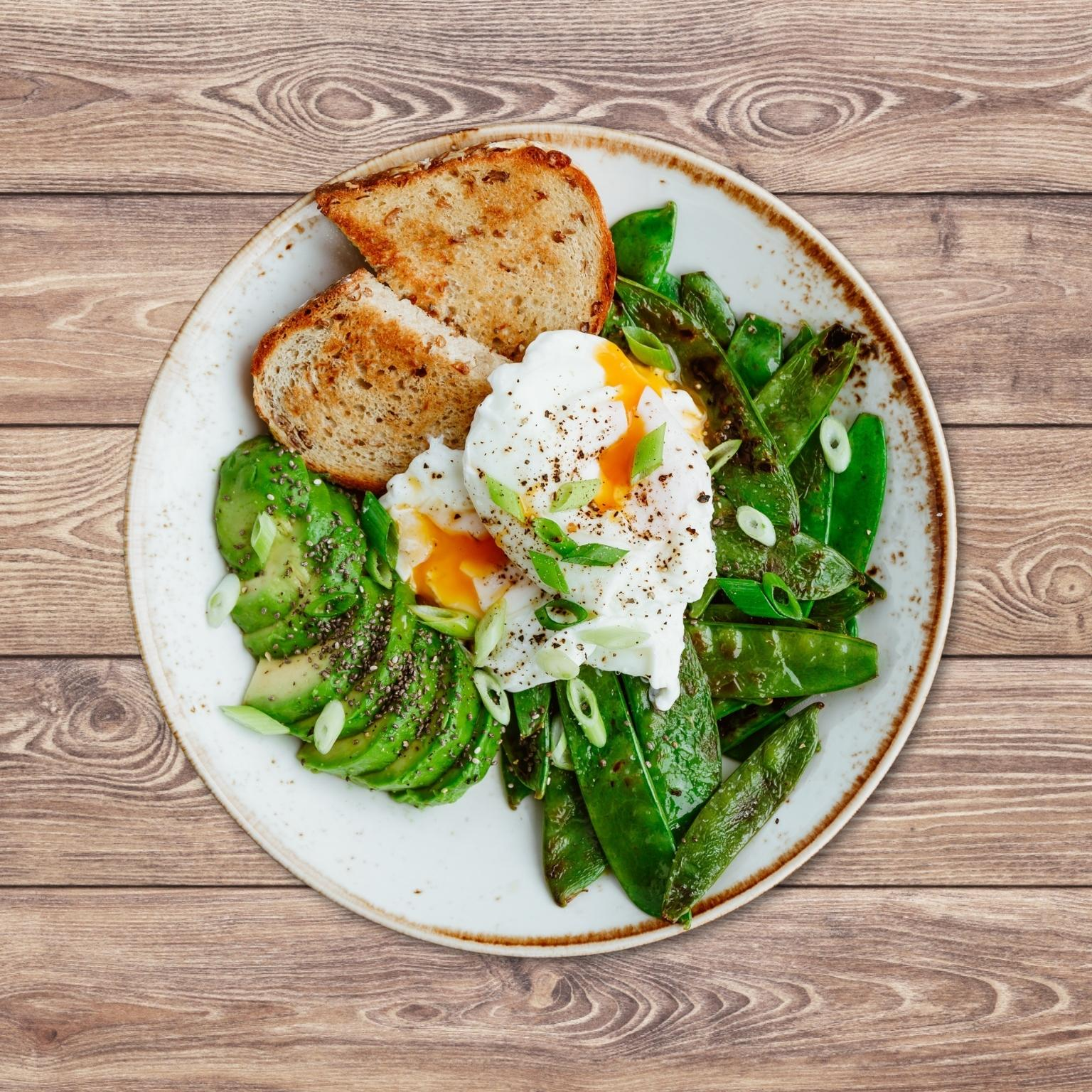 Draufsicht auf einen Teller mit Toast, Avocadoscheiben, pochierten Eiern und Zuckerschoten, auf Holz.