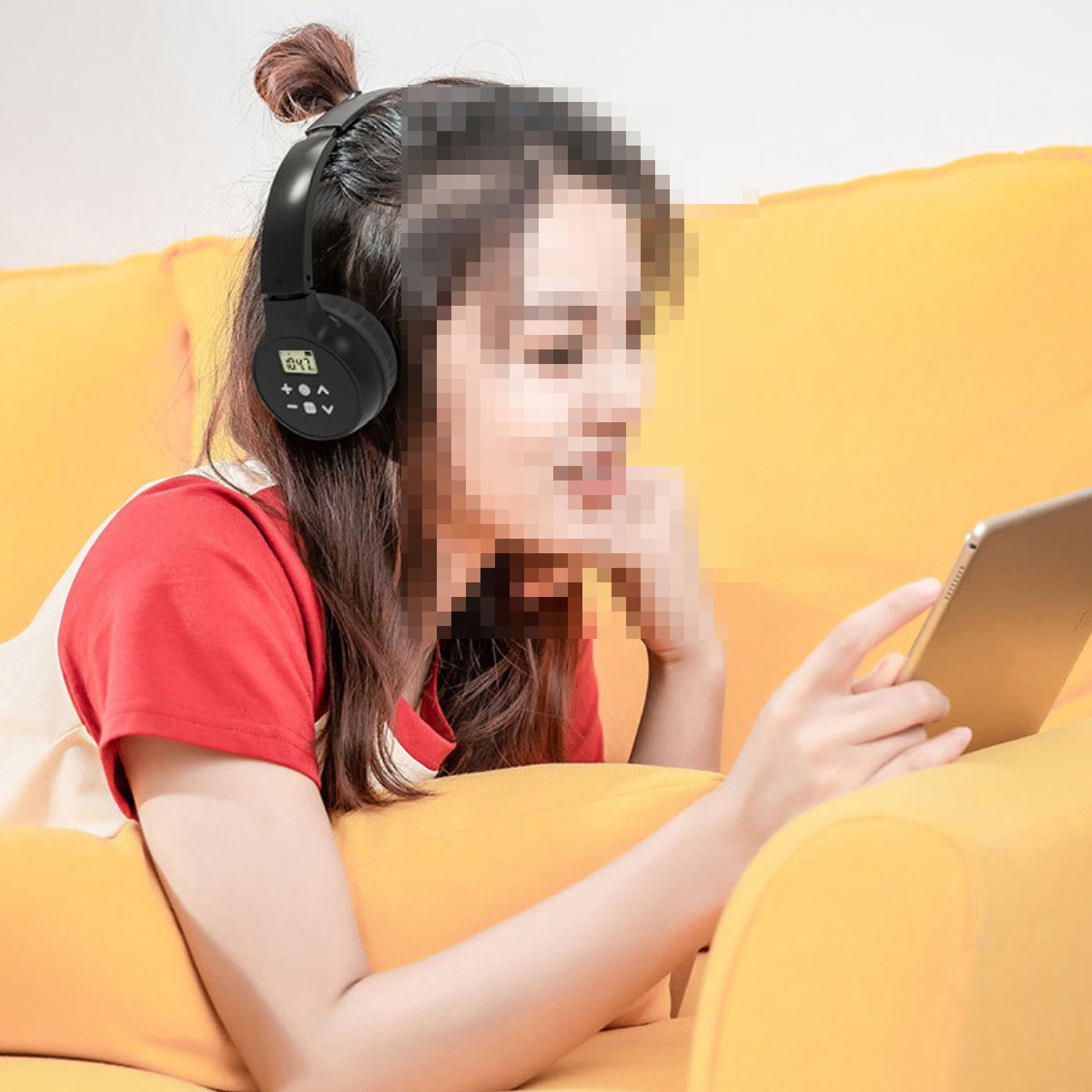 Eine Frau mit schwarzen Kopfhörern, die auf ein Tablet schaut, sitzt auf einem gelben Sofa.