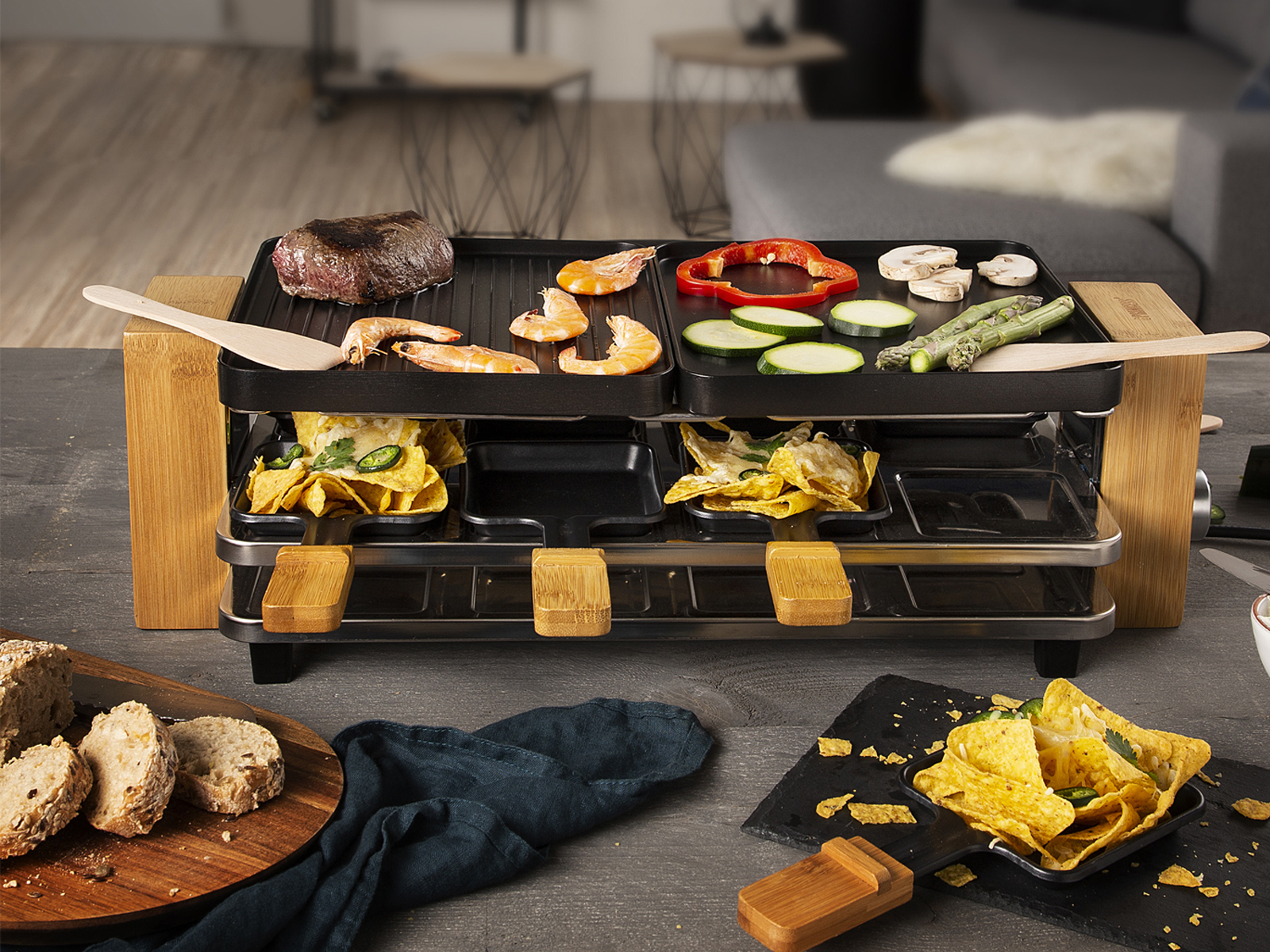 Raclette-Grill mit verschiedenen Speisen und Holzspateln auf einem Tisch.