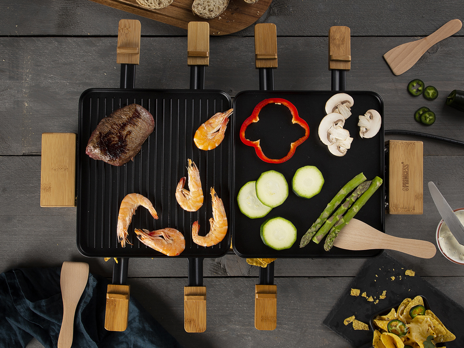 Raclette-Grill mit Fleisch, Garnelen und Gemüse auf einem Holztisch.