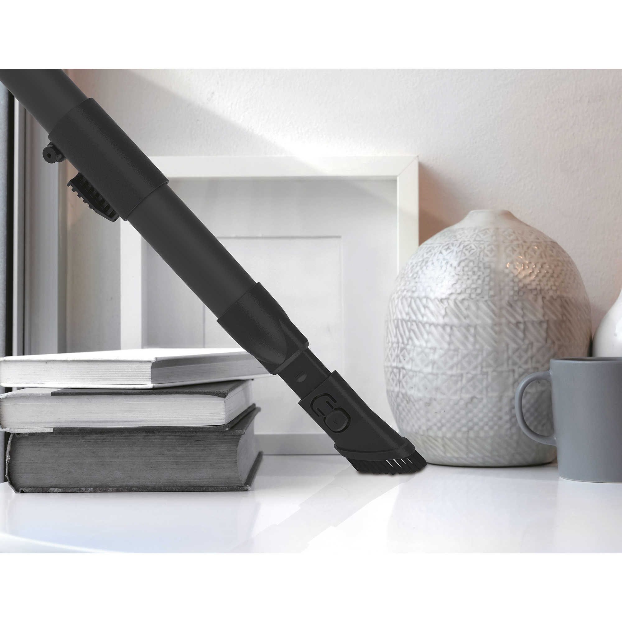 Zbliżenie końcówki odkurzacza czyszczącej białą powierzchnię, z wazonem i książkami w tle.